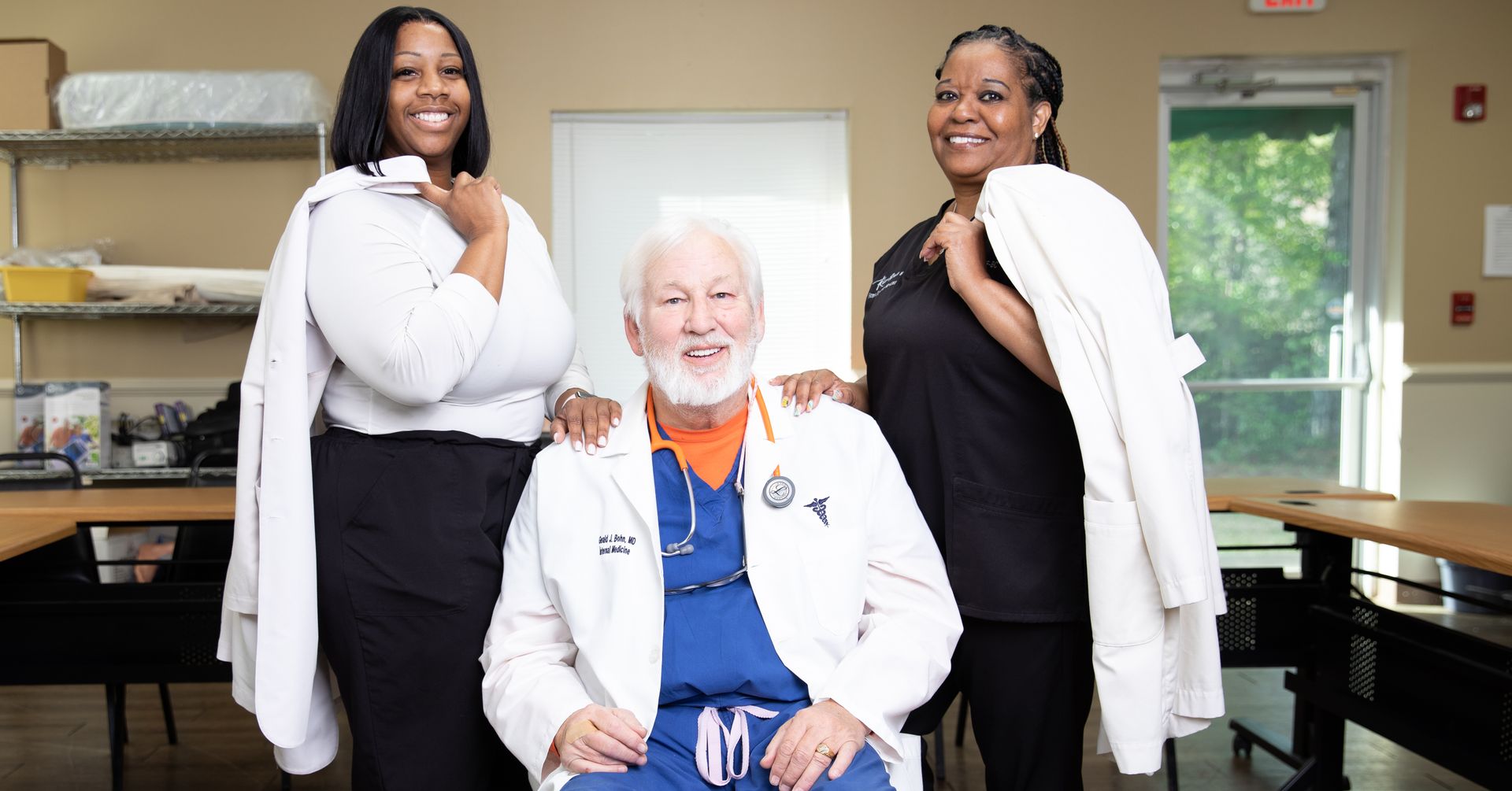 A man in a lab coat is sitting in a chair with two nurses standing behind him.