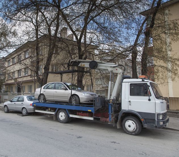 A white tow truck carries a silver car on its flatbed, parked on a street in front of a building and trees.