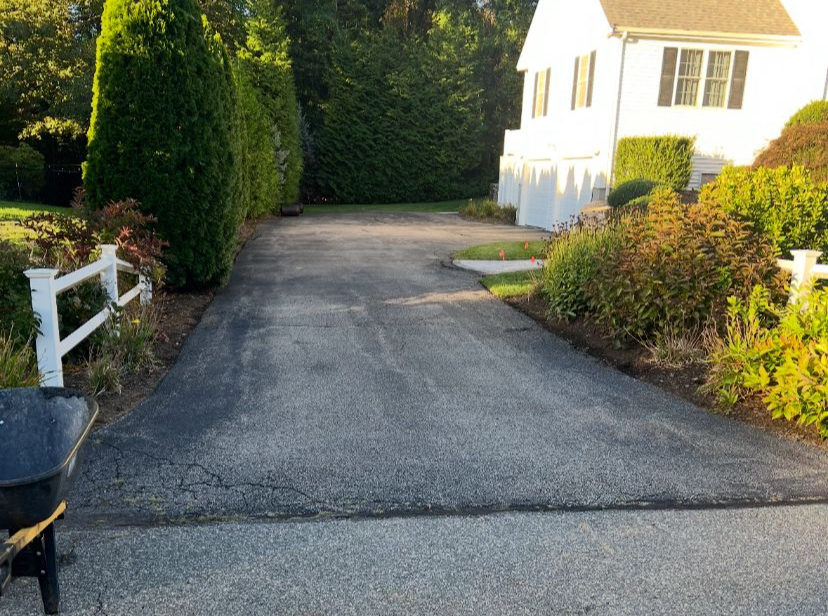 Asphalt driveway leading to a white house, bordered by bushes, trees, and a white fence.