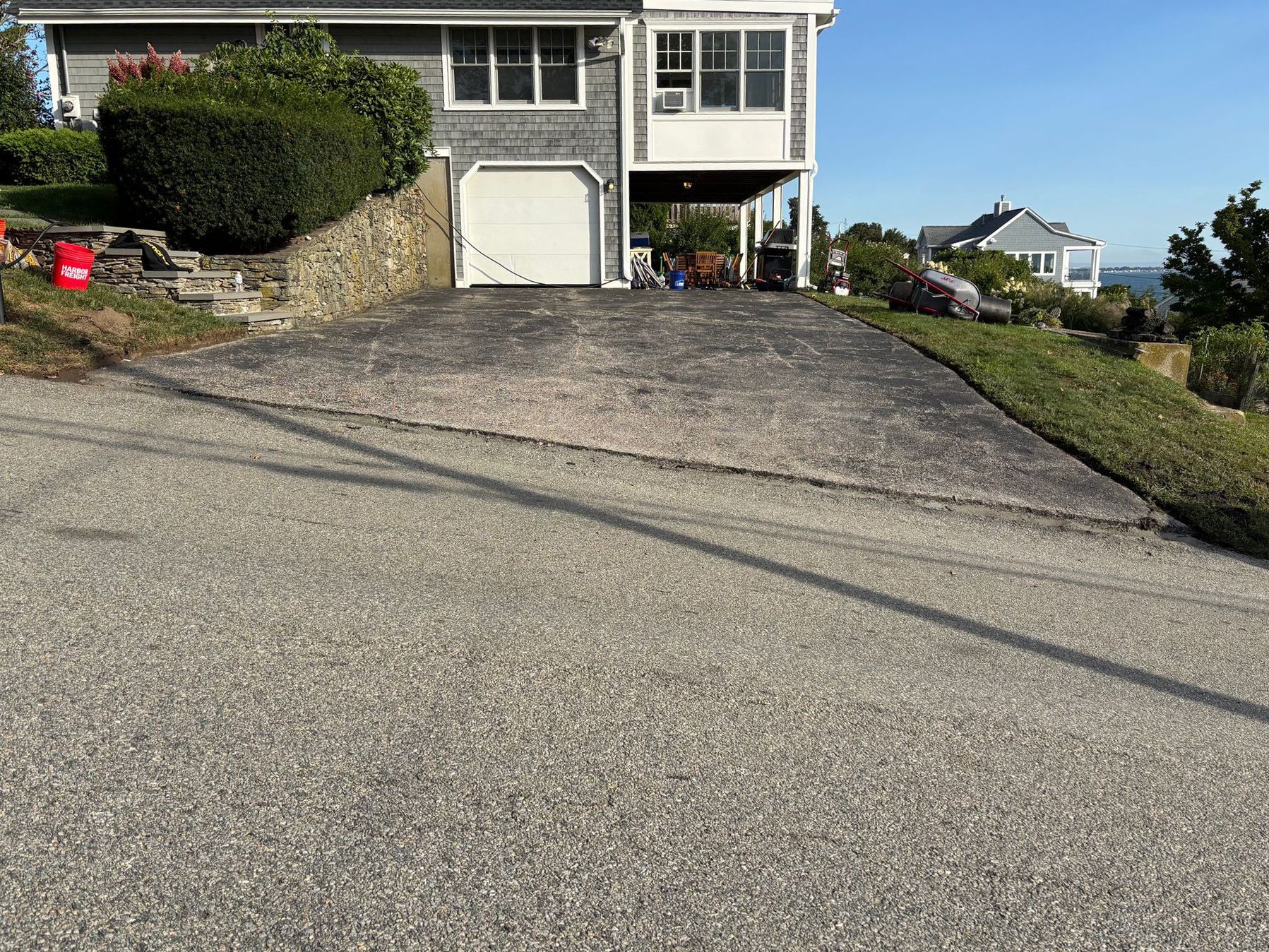Asphalt driveway leading up to a gray house with a garage; a grassy slope is to the right.