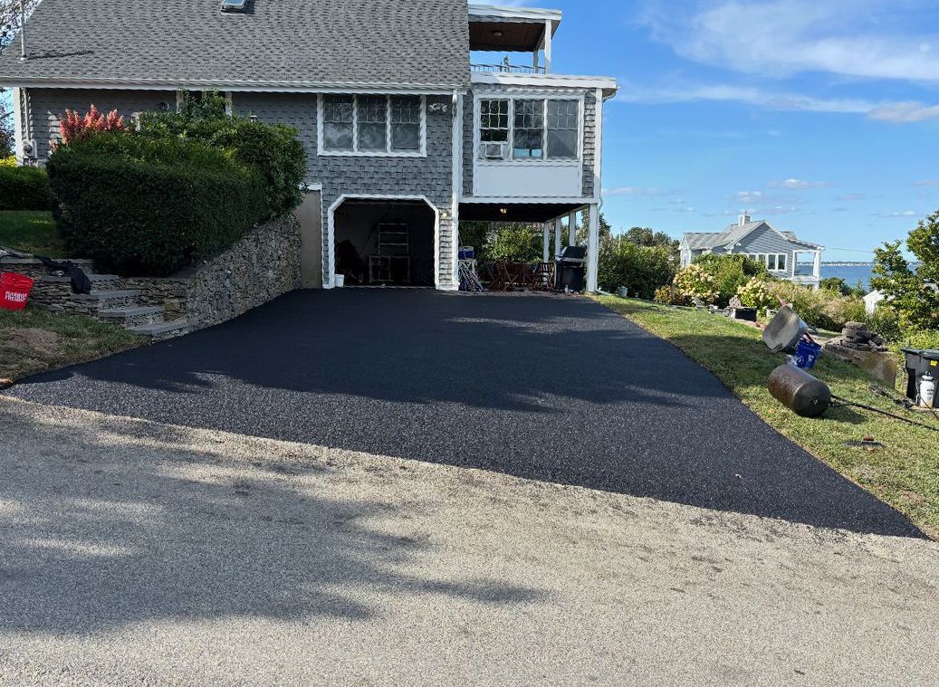 Newly paved blacktop driveway leading to a gray house with a garage and a view of the ocean.