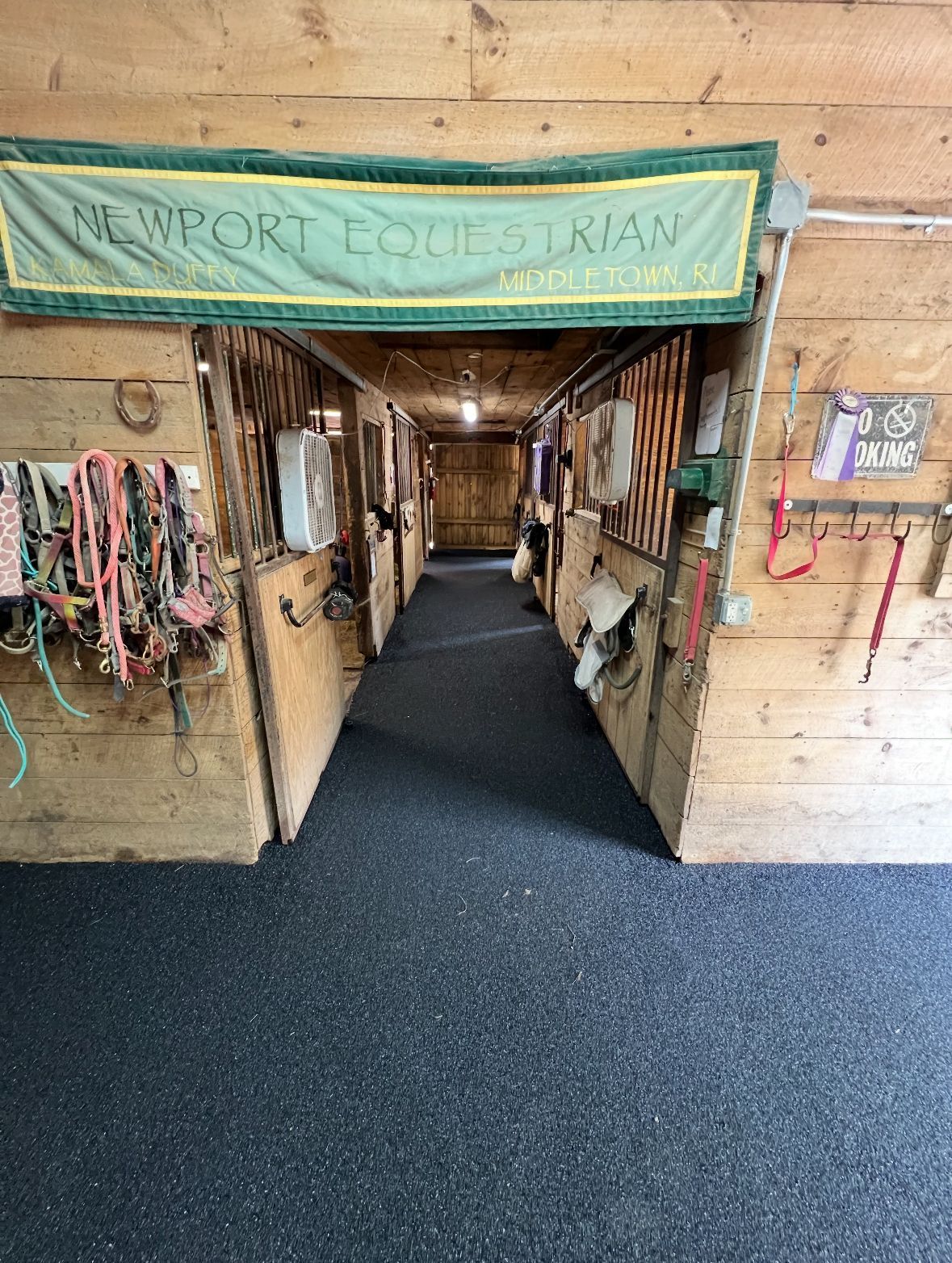 A barn aisle with horse stalls, tack, and a sign that says Newport Equestrian.