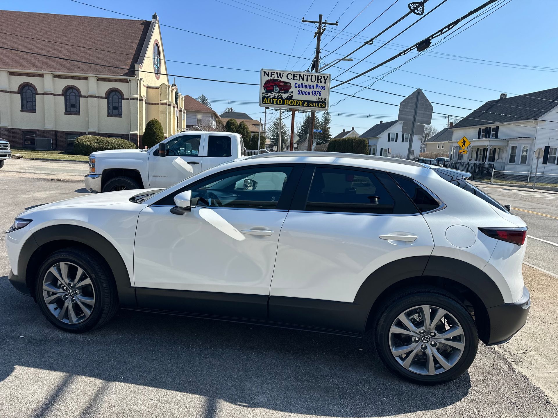 A side view of a white Mazda CX-30 parked on an asphalt lot near a church and a suburban street.