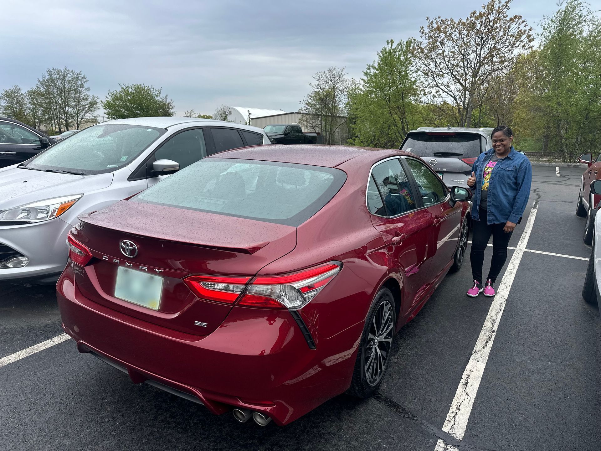 Maroon sedan parked in a lot beside a woman in a blue jacket, with gray cars and trees in the background