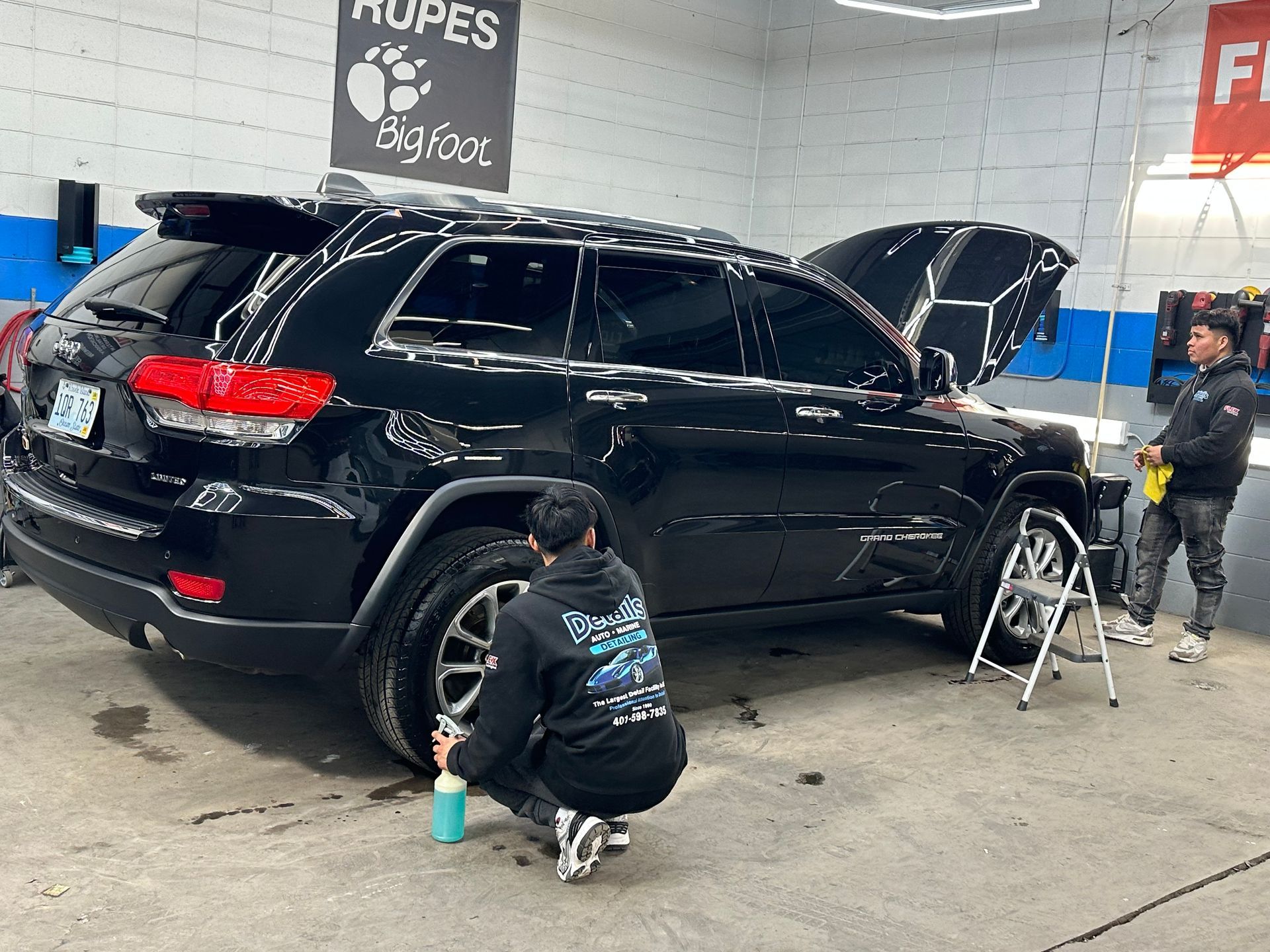 People are cleaning a black SUV in a garage. One sprays a tire, the other wipes the car.