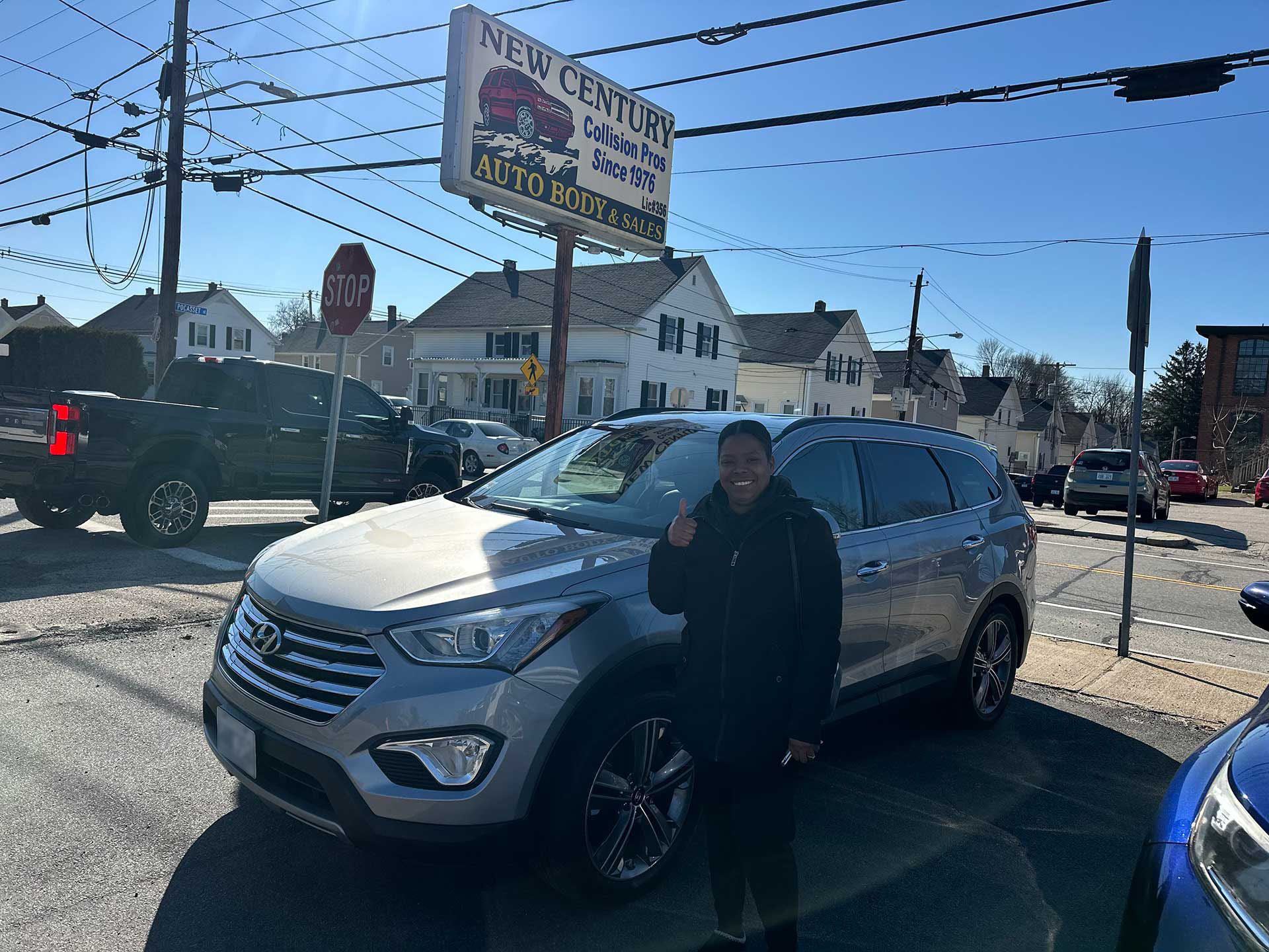 A person smiling while standing next to a silver SUV at a car dealership on a sunny day.