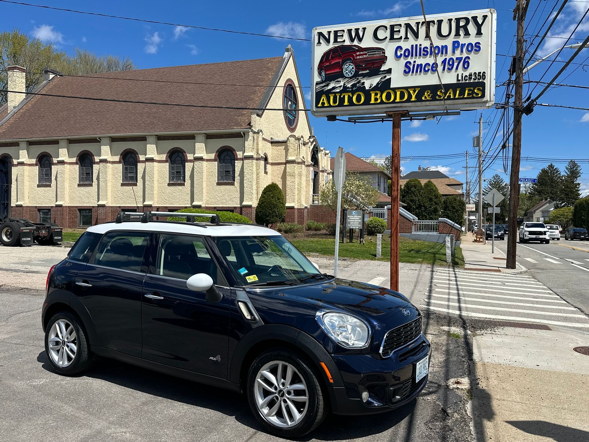 Dark blue Mini Cooper parked beside a New Century sign outside a stone church on a sunny street.
