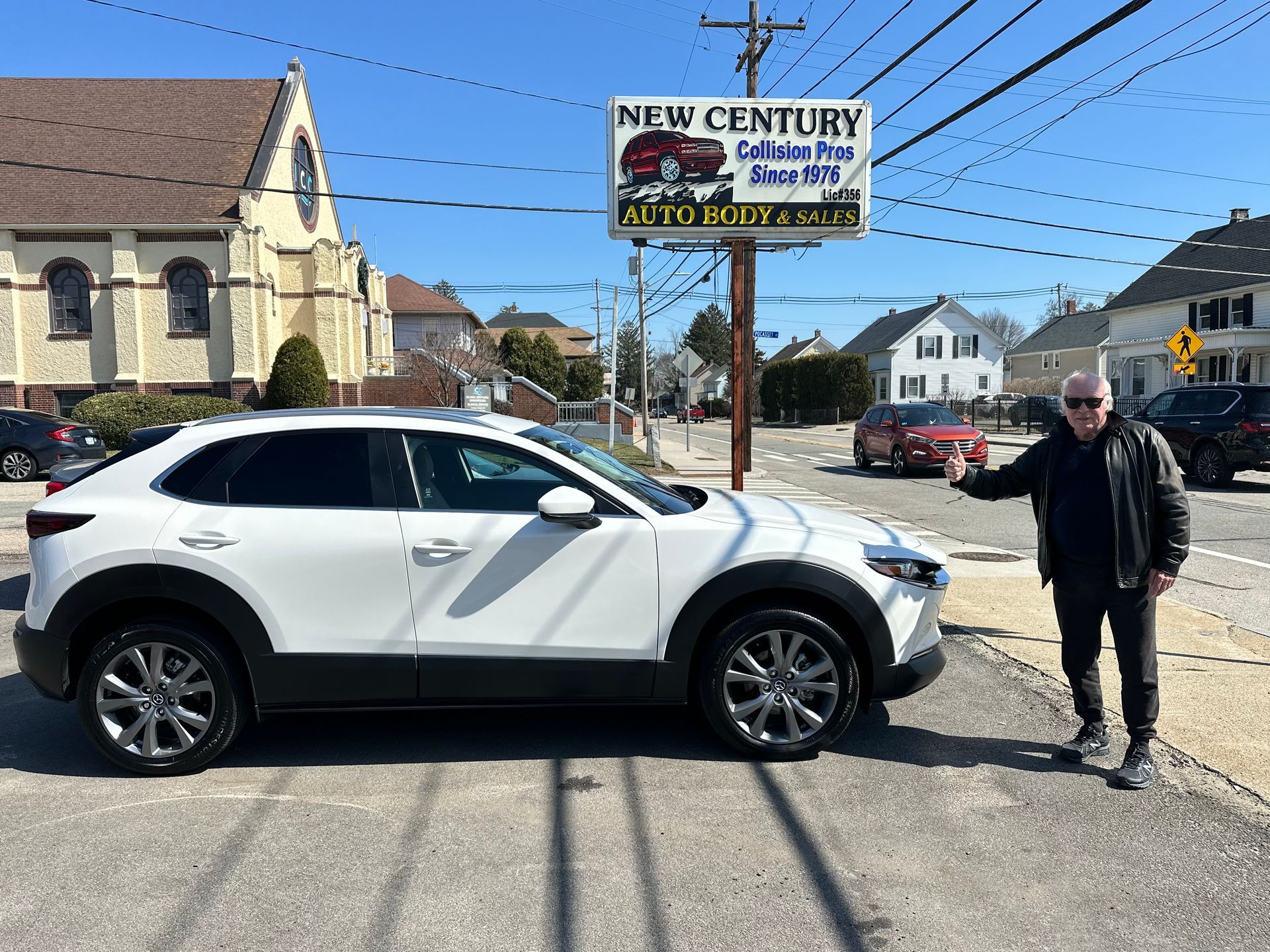 A person in a black jacket stands next to a white SUV parked in front of a New Century Auto Body shop on a sunny day.