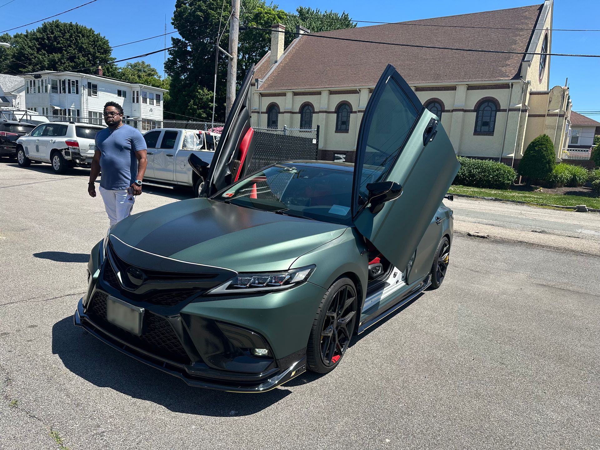 Matte green sports car with gullwing door open on a street, man standing nearby, church in background