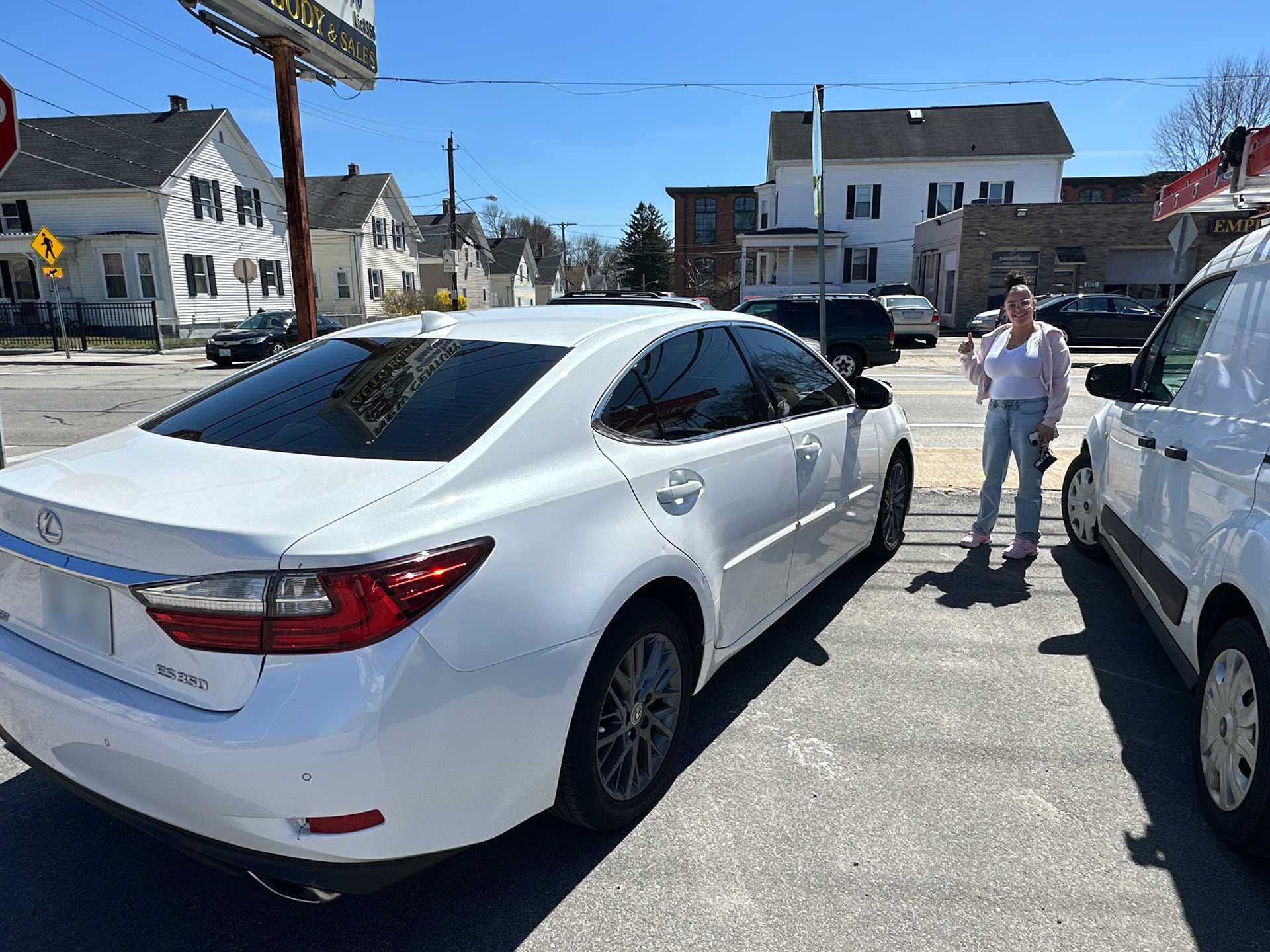 A person stands next to a white sedan parked on a sunny street near residential buildings and a commercial sign.