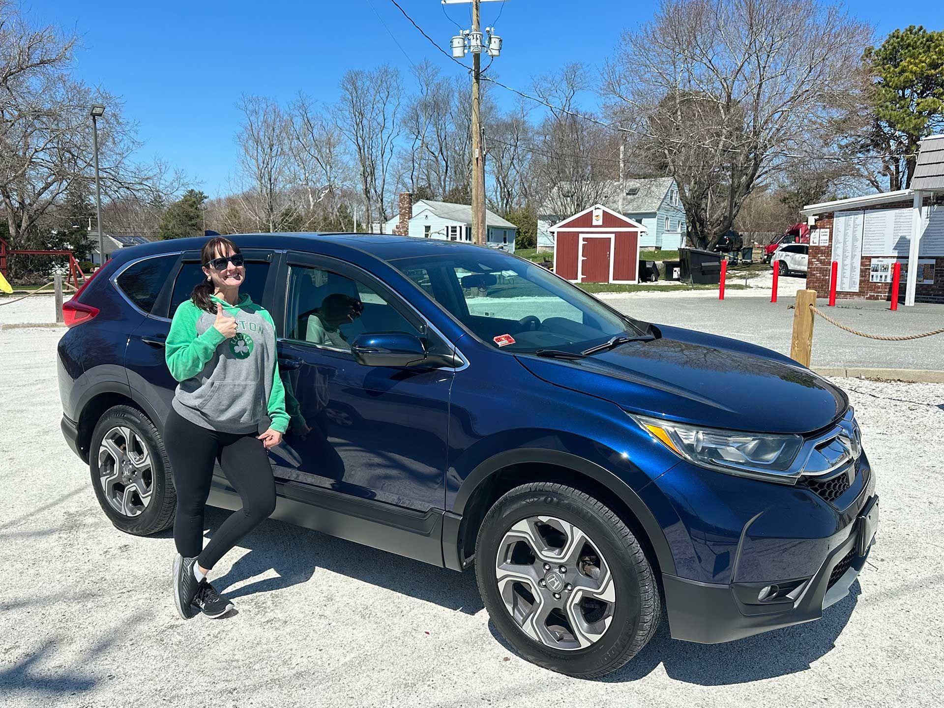 A person in a green hoodie stands on a gravel lot posing next to a dark blue SUV on a sunny day.