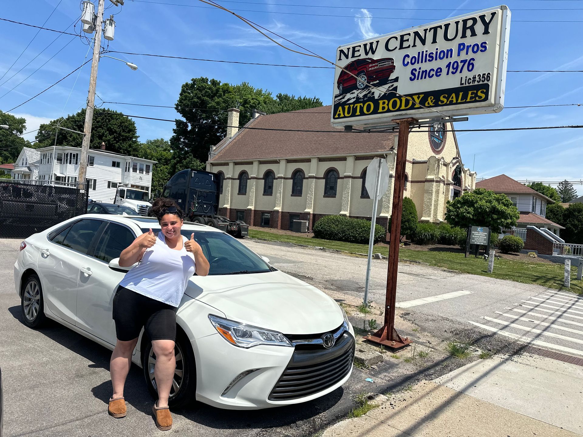 Person leaning on a white car at a used auto body sales lot with a large sign overhead.