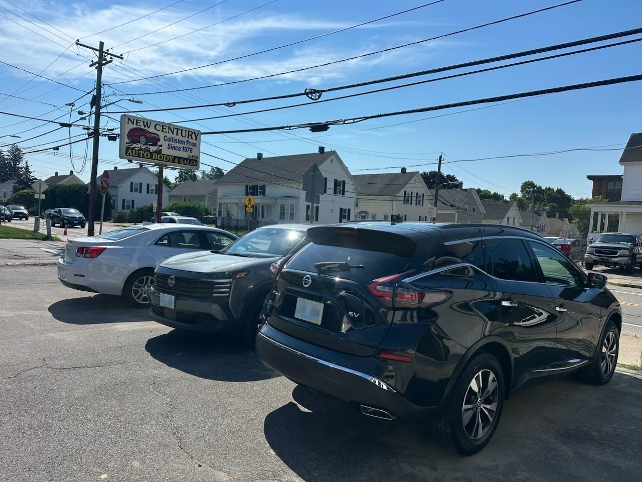 Three parked cars in a sunny parking lot with houses, power lines, and a roadside sign in the background