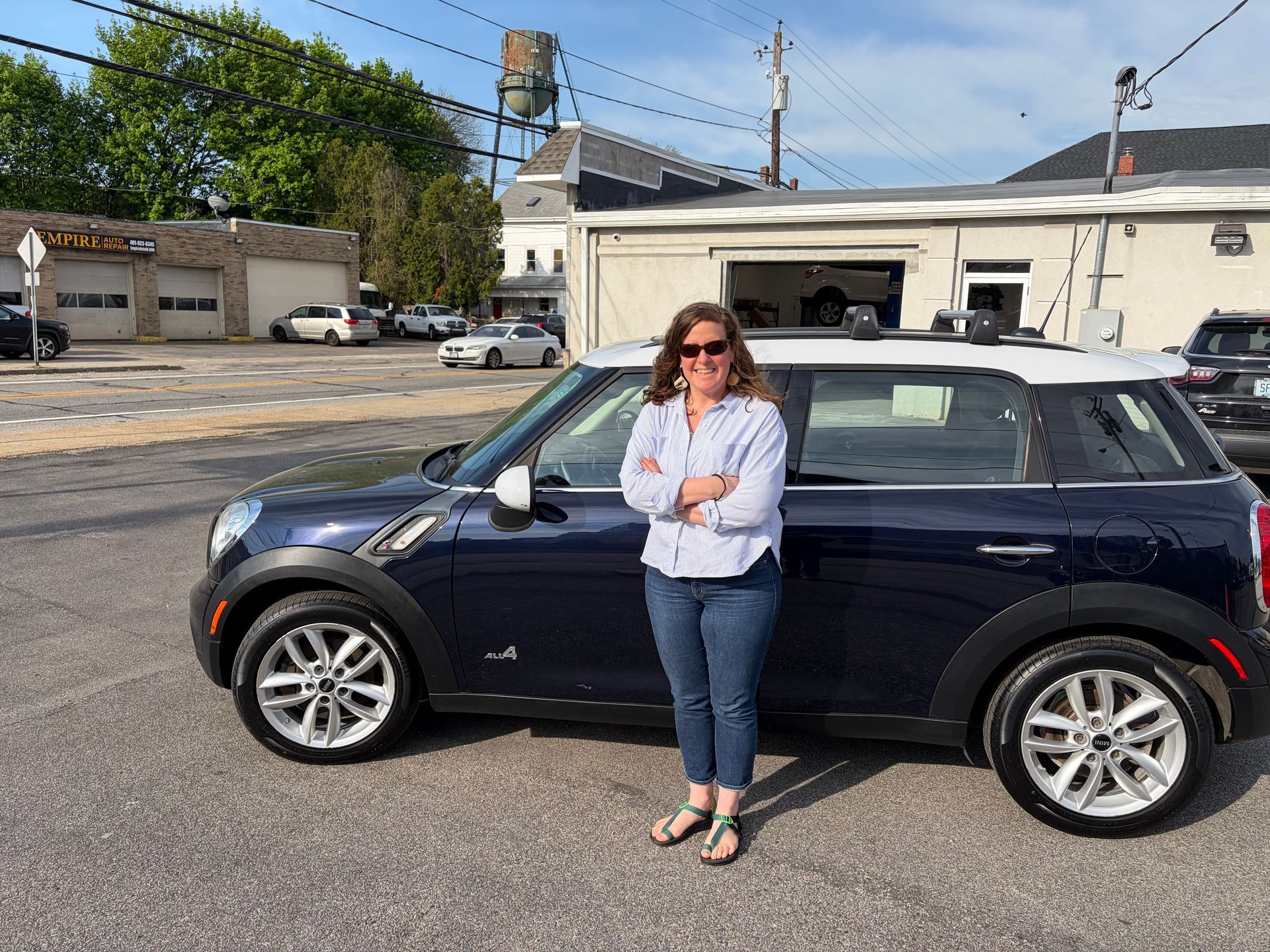 Woman standing beside a dark blue Mini Cooper in a parking lot, arms crossed, wearing sunglasses.