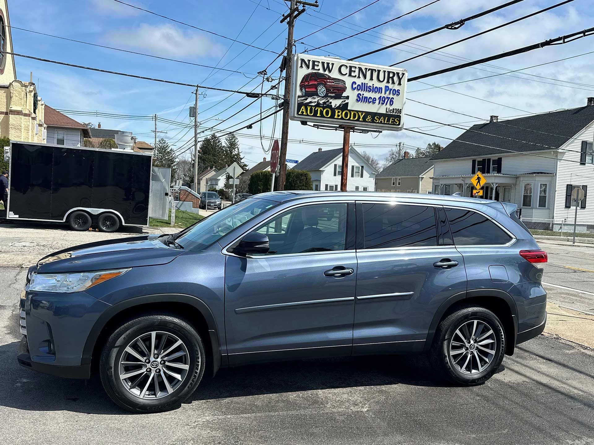 A dark gray SUV parked on a sunny day in front of a New Century Auto sales lot with a sign and nearby residential houses.