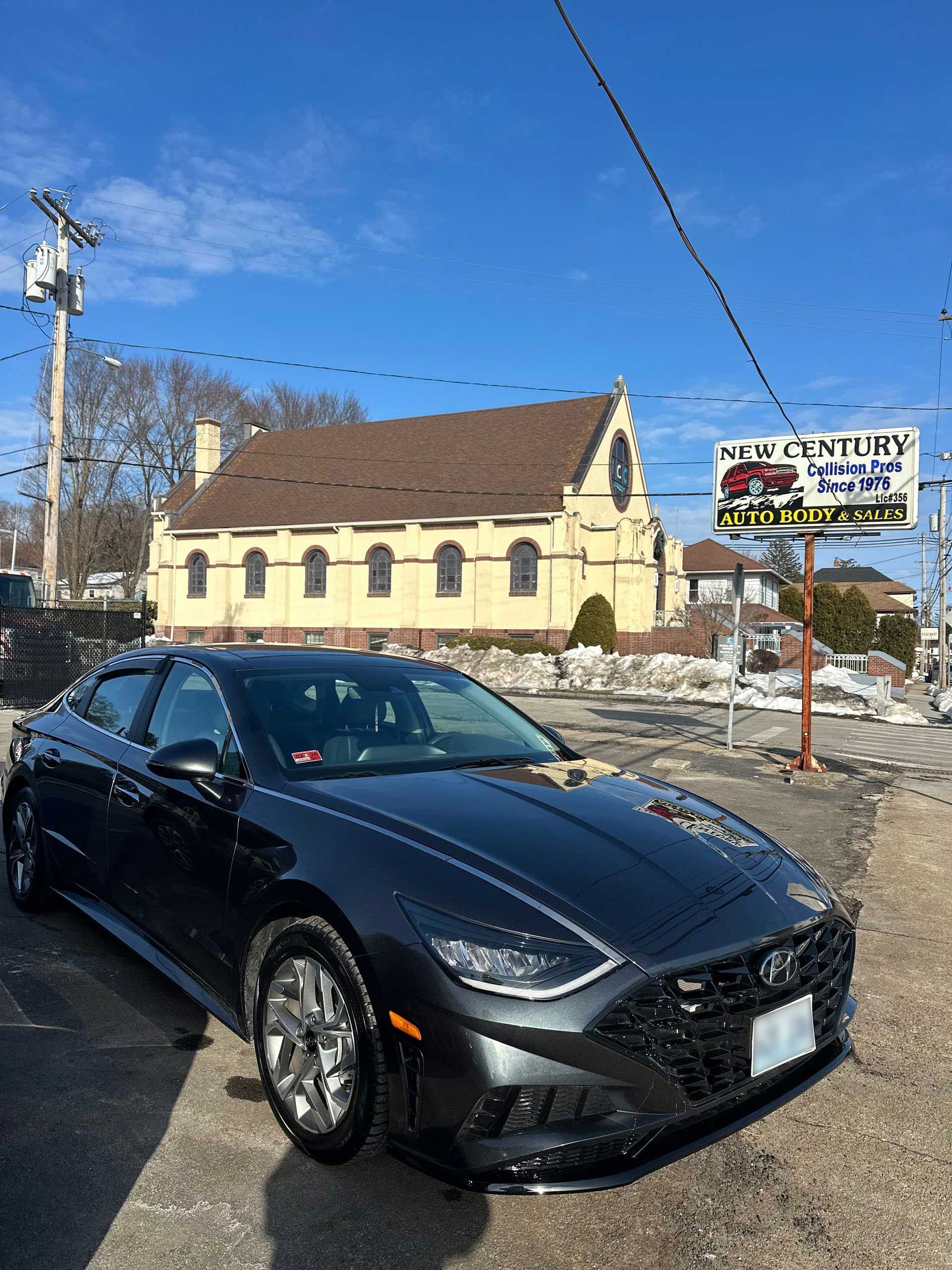 A dark gray sedan parked on a gravel lot in front of a tan stone church under a bright blue sky.