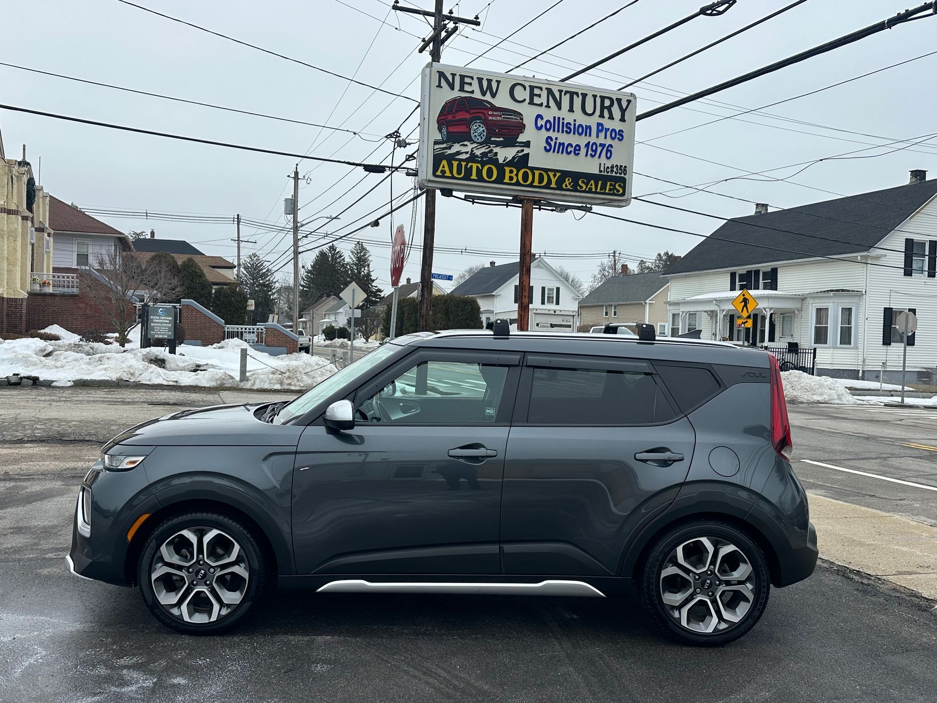 Gray Kia Soul parked in front of New Century Collision auto body shop on a cloudy day.