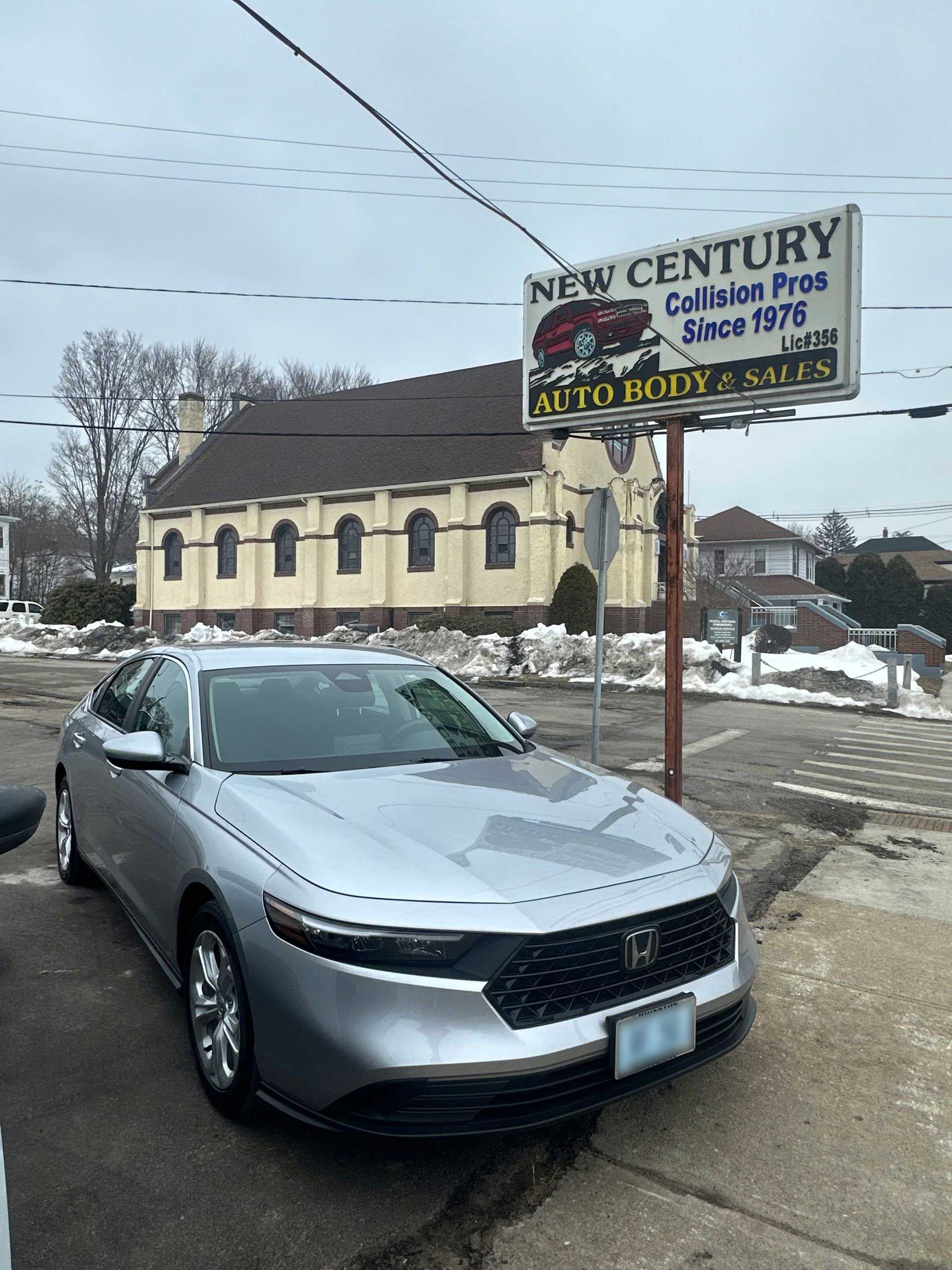 A silver Honda sedan parked in front of a New Century Auto Body sign with a large church building in the background.