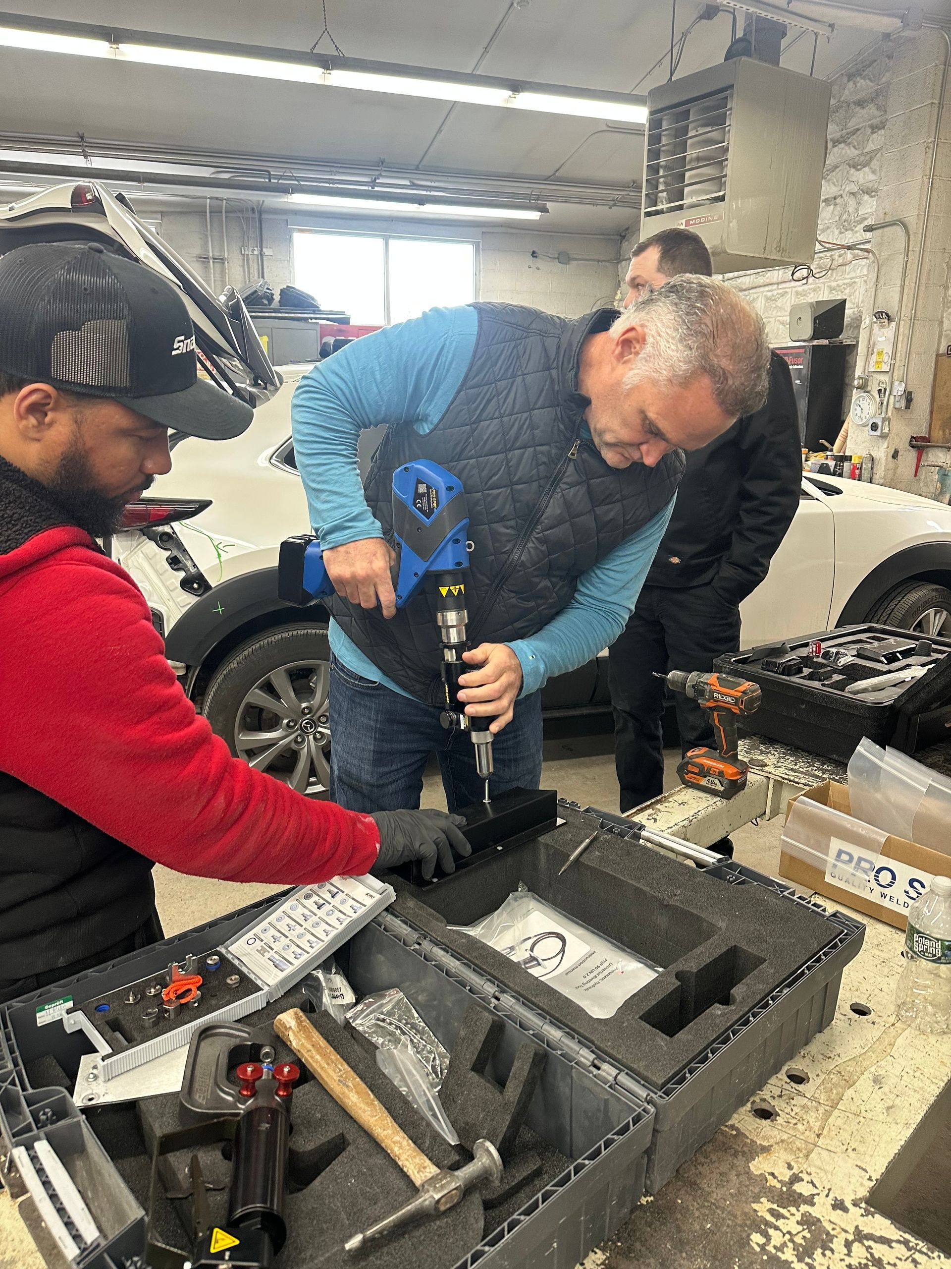 Three people work on equipment at a workbench. One holds a drill, another looks on, and a third works from a toolbox.