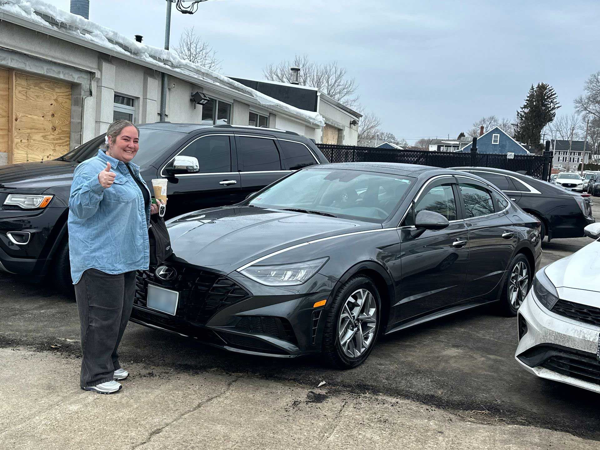 A person smiling and giving a thumbs up next to a gray sedan in a car dealership parking lot.