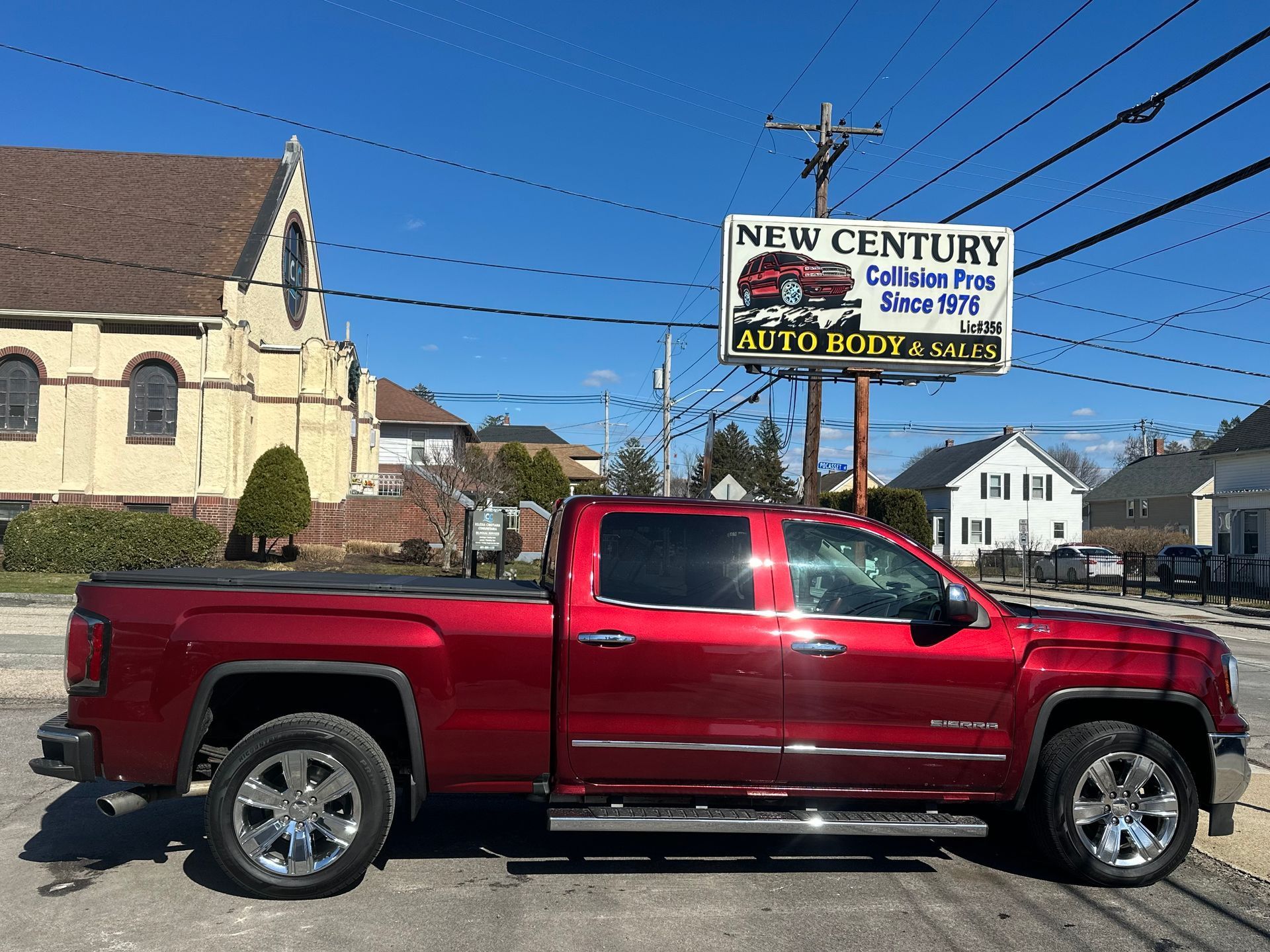A side view of a shiny red pickup truck parked in front of a business sign for New Century Auto Body near a church.