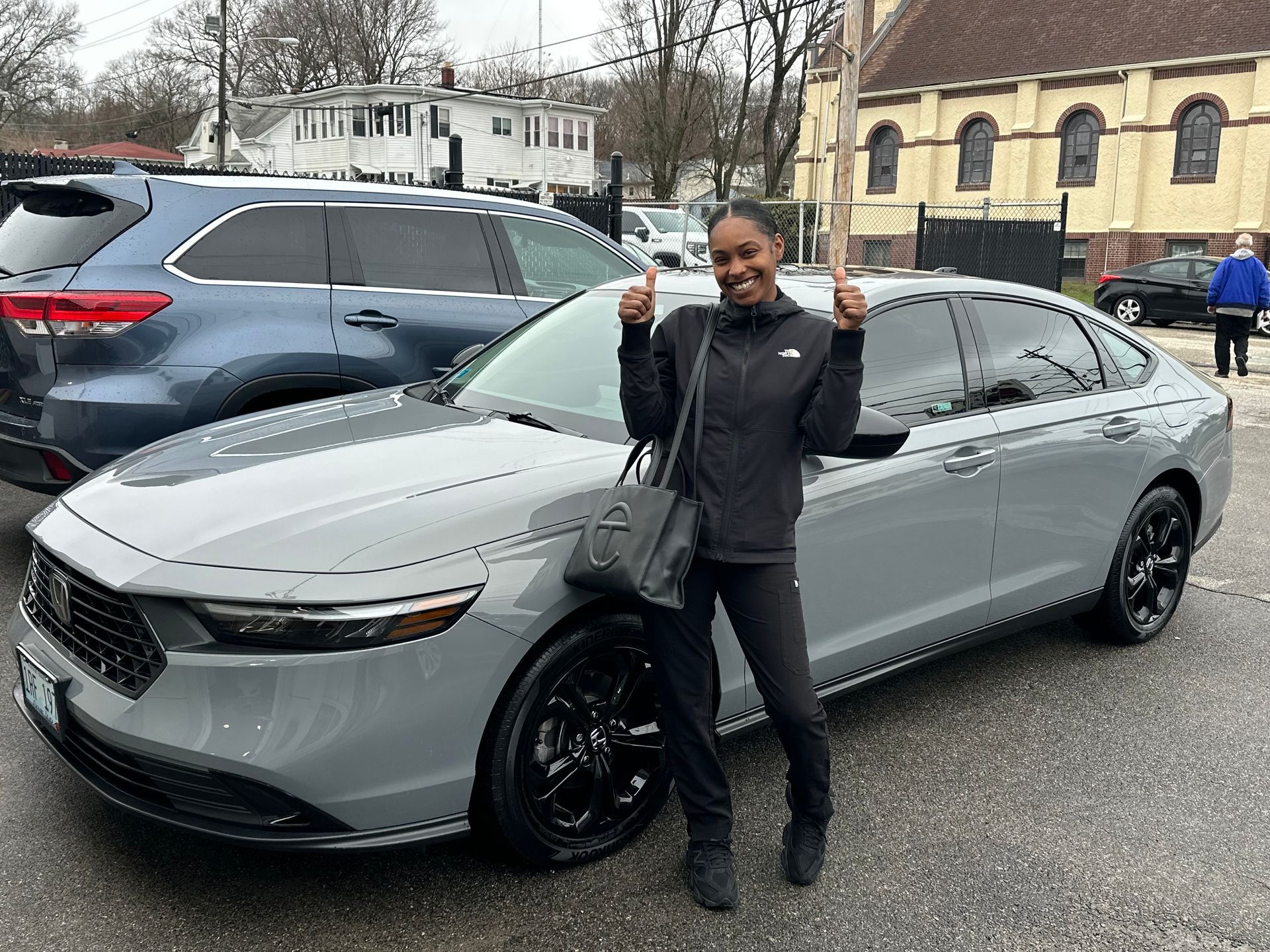 A smiling person standing with both thumbs up in front of a gray sedan in an outdoor parking lot near a church building.