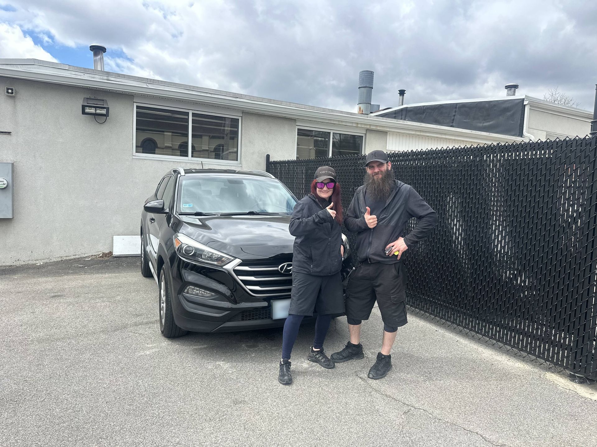 Two people posing beside a black SUV in a driveway beside a white house under cloudy skies