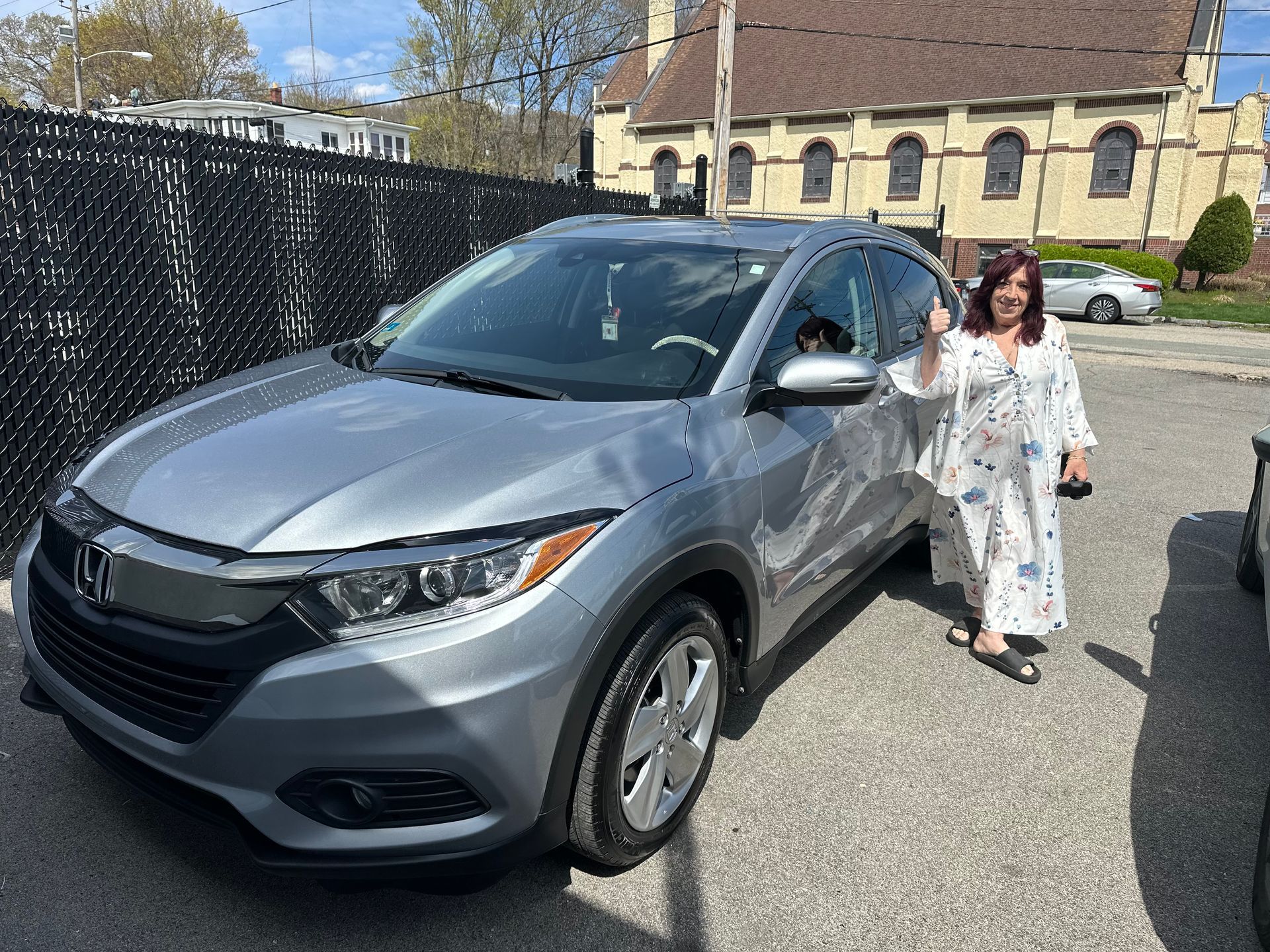 Woman standing beside a silver SUV in a parking lot on a sunny day.