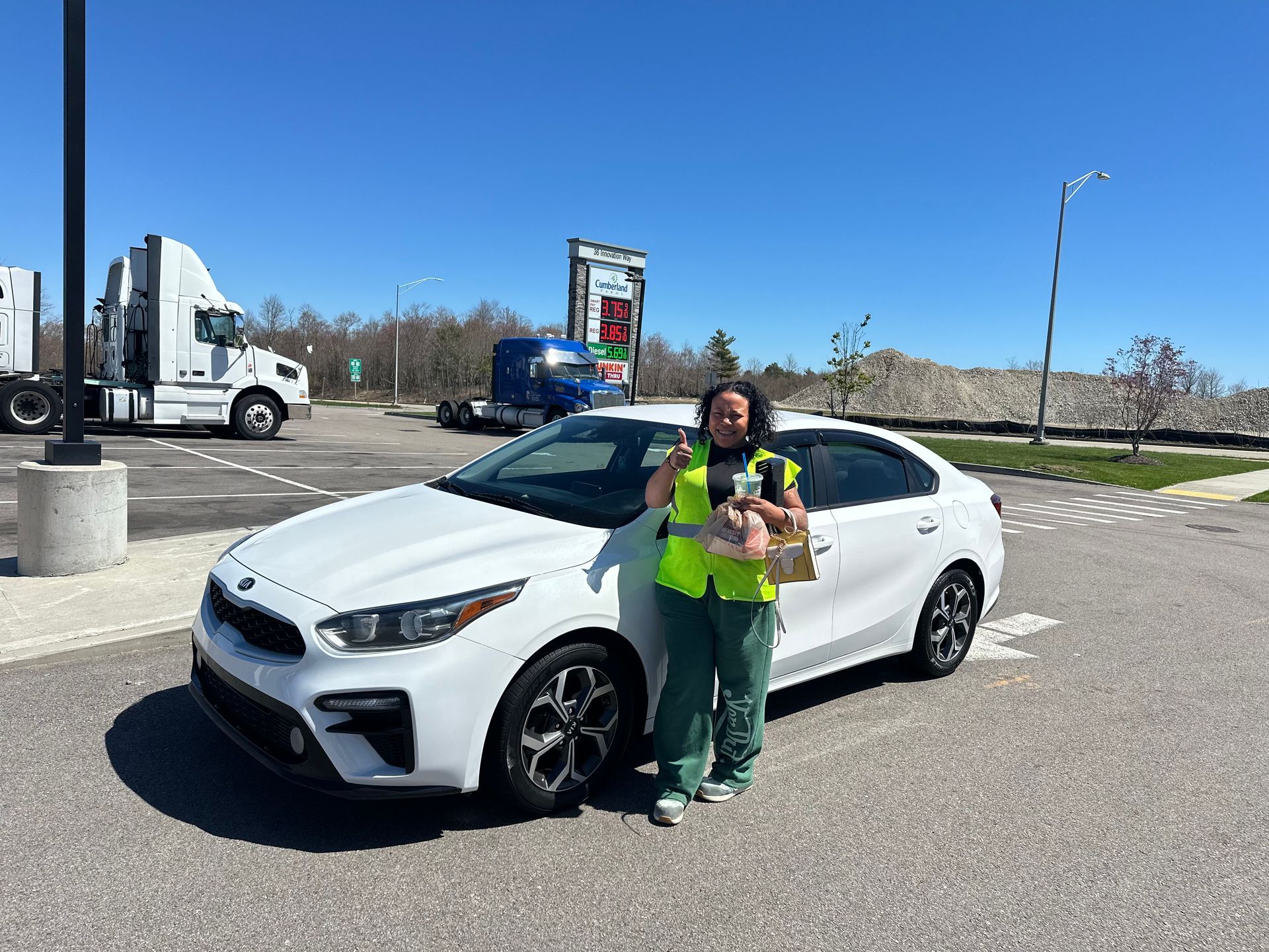 Woman holding a dog beside a white sedan in a parking lot on a sunny day. 