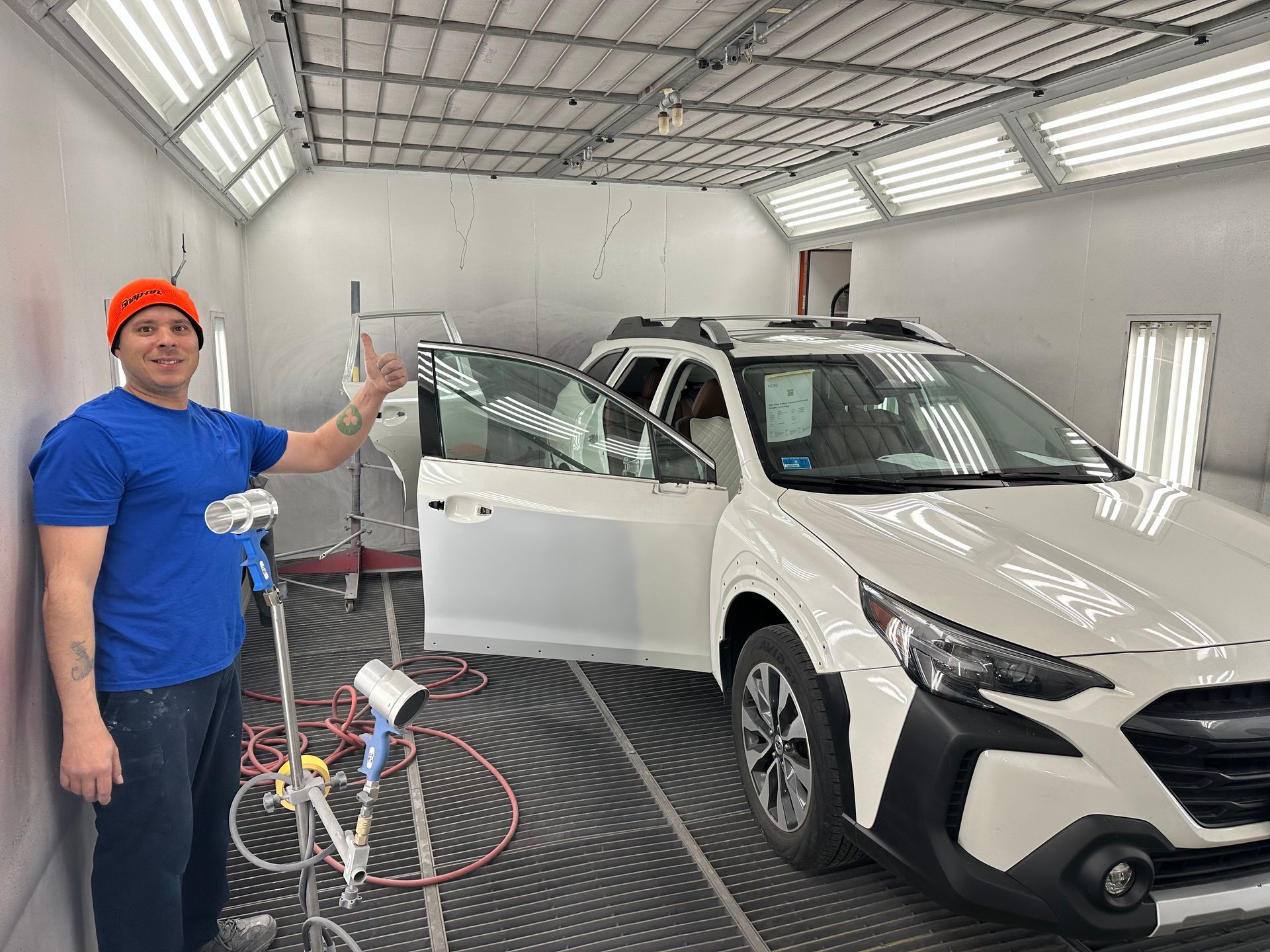 Man in blue shirt with spray gun beside white SUV in auto body paint booth. 