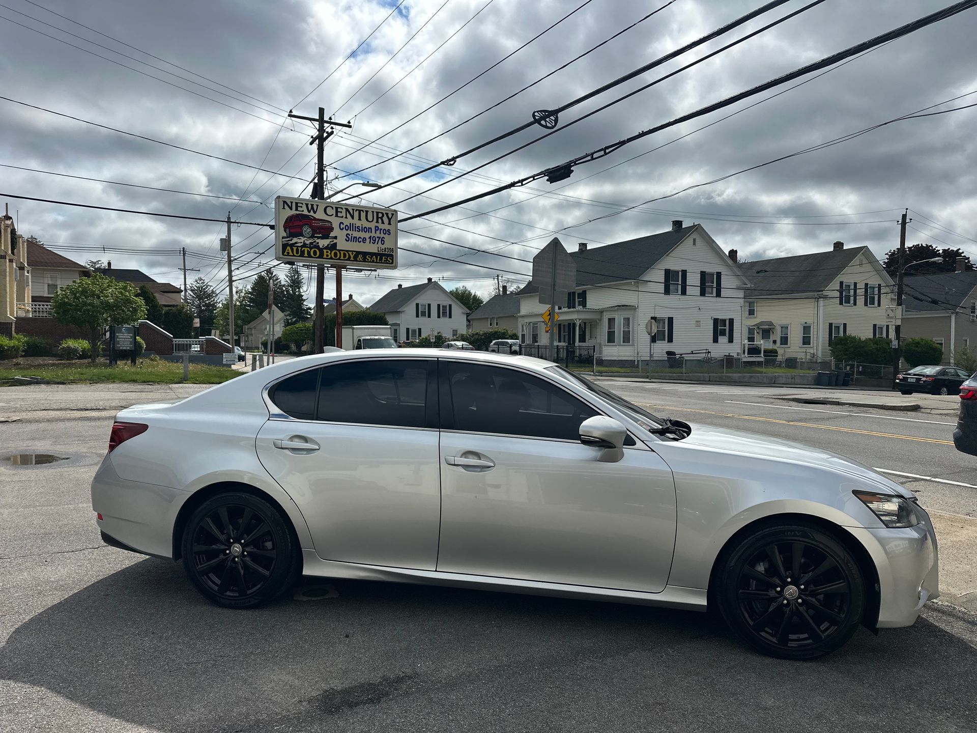White sedan parked on a roadside under power lines, with houses and a cloudy sky in the background