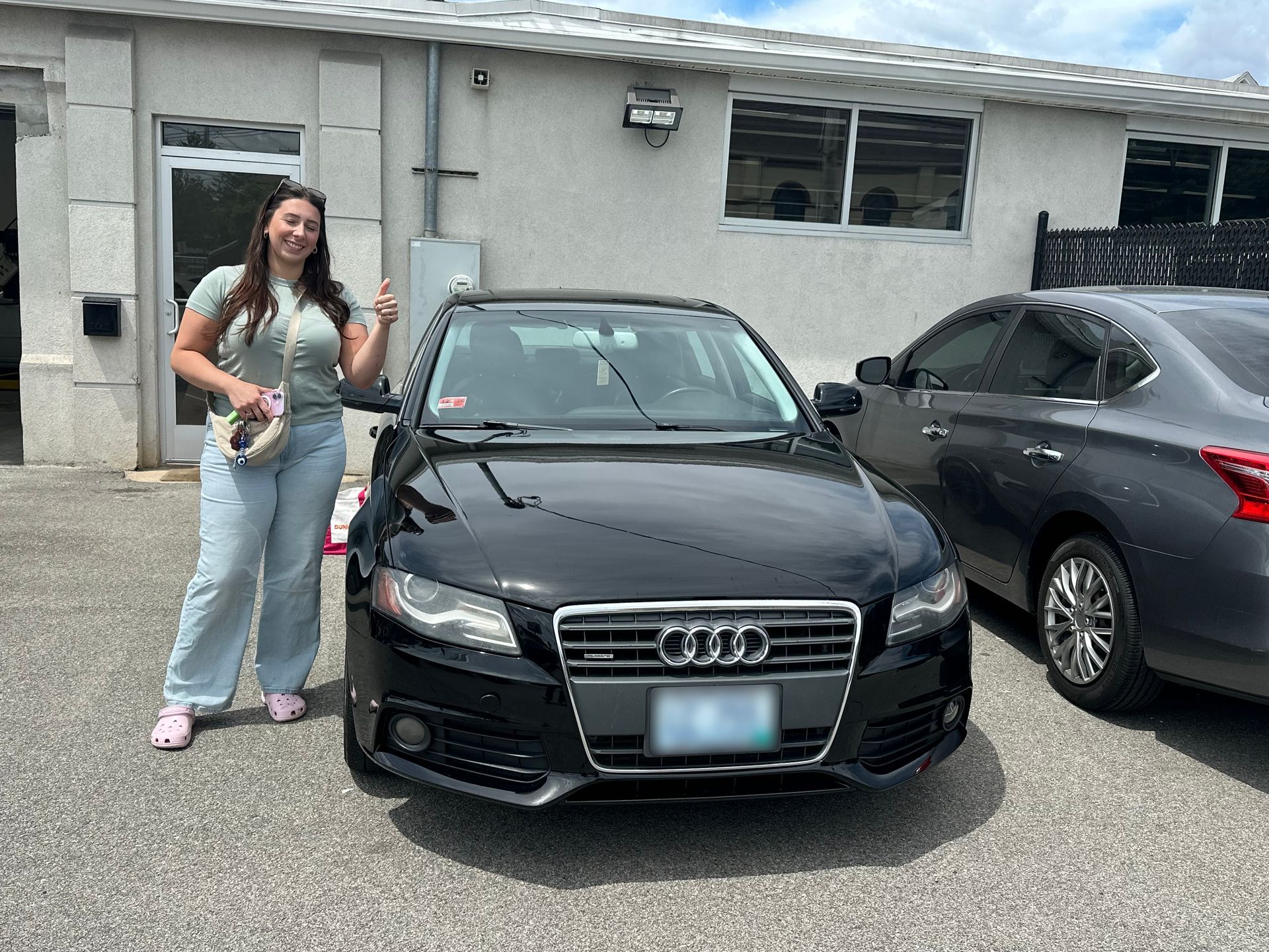Woman stands beside a black Audi sedan in a parking lot, smiling and giving a thumbs-up.