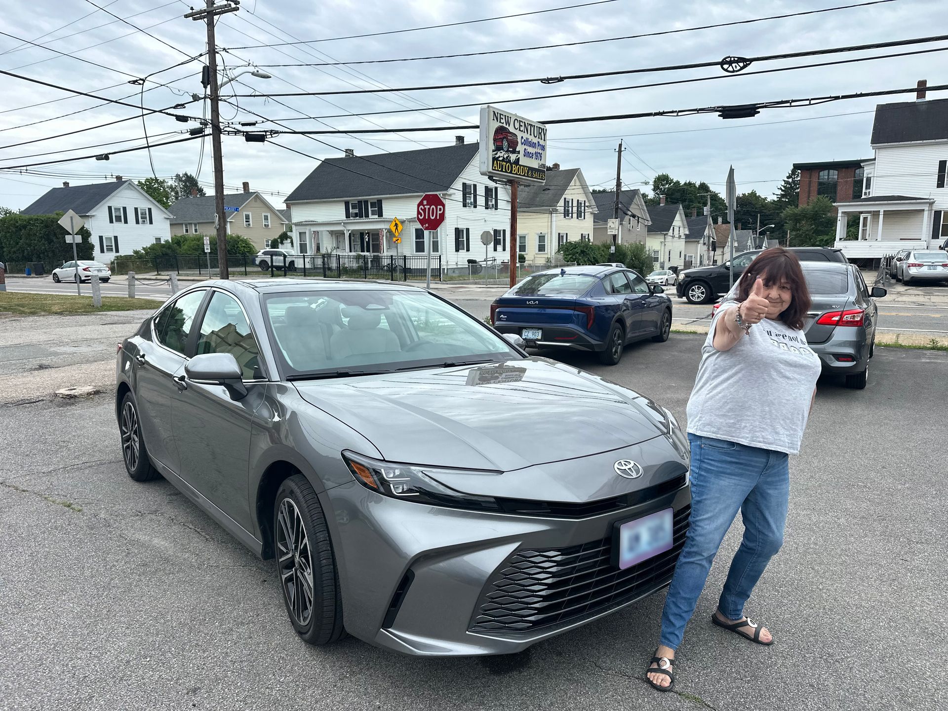 A gray sedan was parked on a street with a woman posing beside it in a small town intersection.