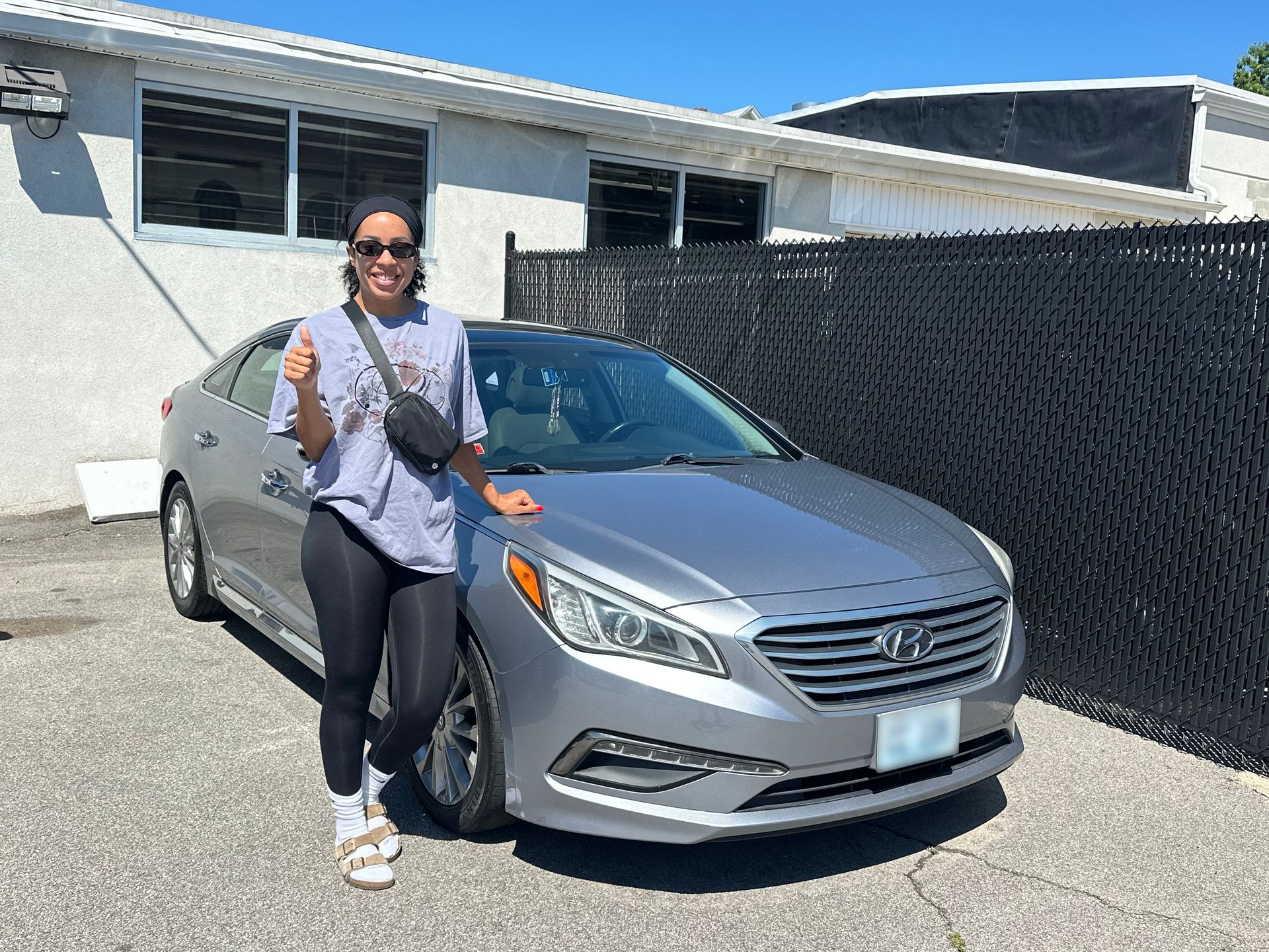 Person standing beside a silver sedan in a sunny driveway, holding a drink and smiling.