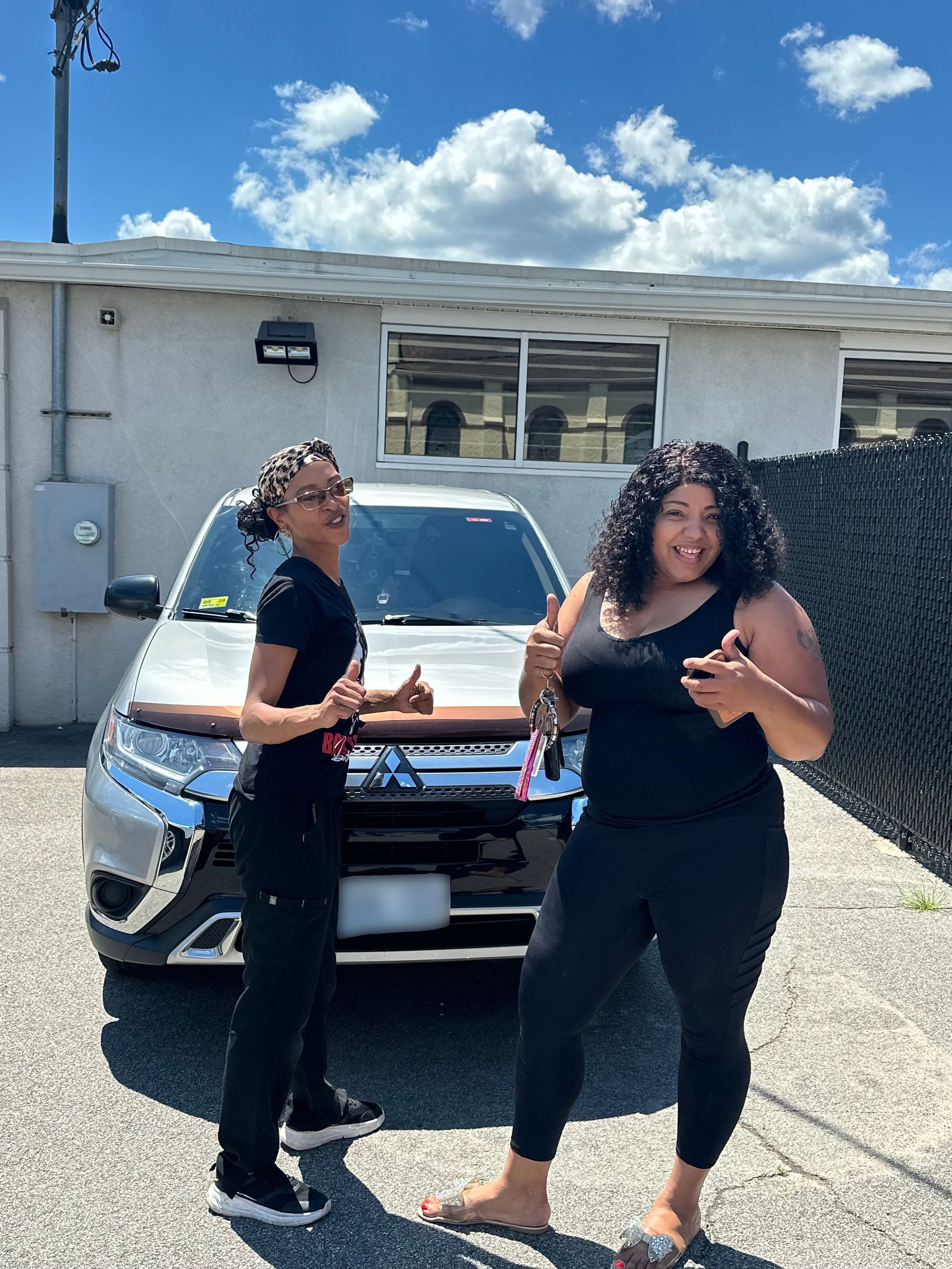 Two people standing in front of a silver SUV in a driveway, smiling in bright sunlight.