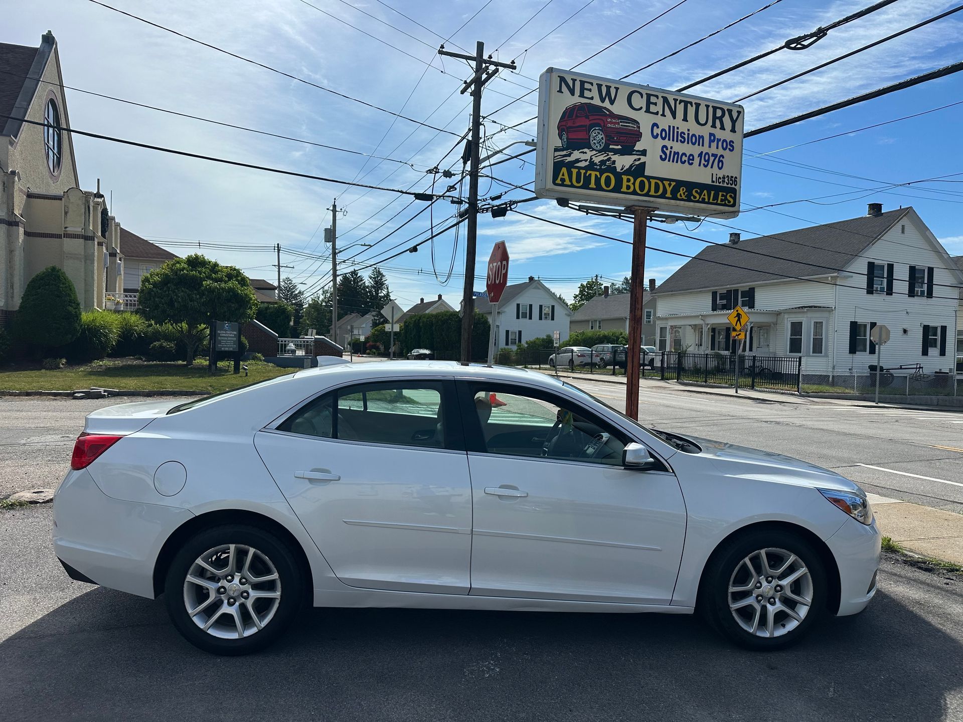 White sedan parked by a roadside sign and buildings on a sunny street.