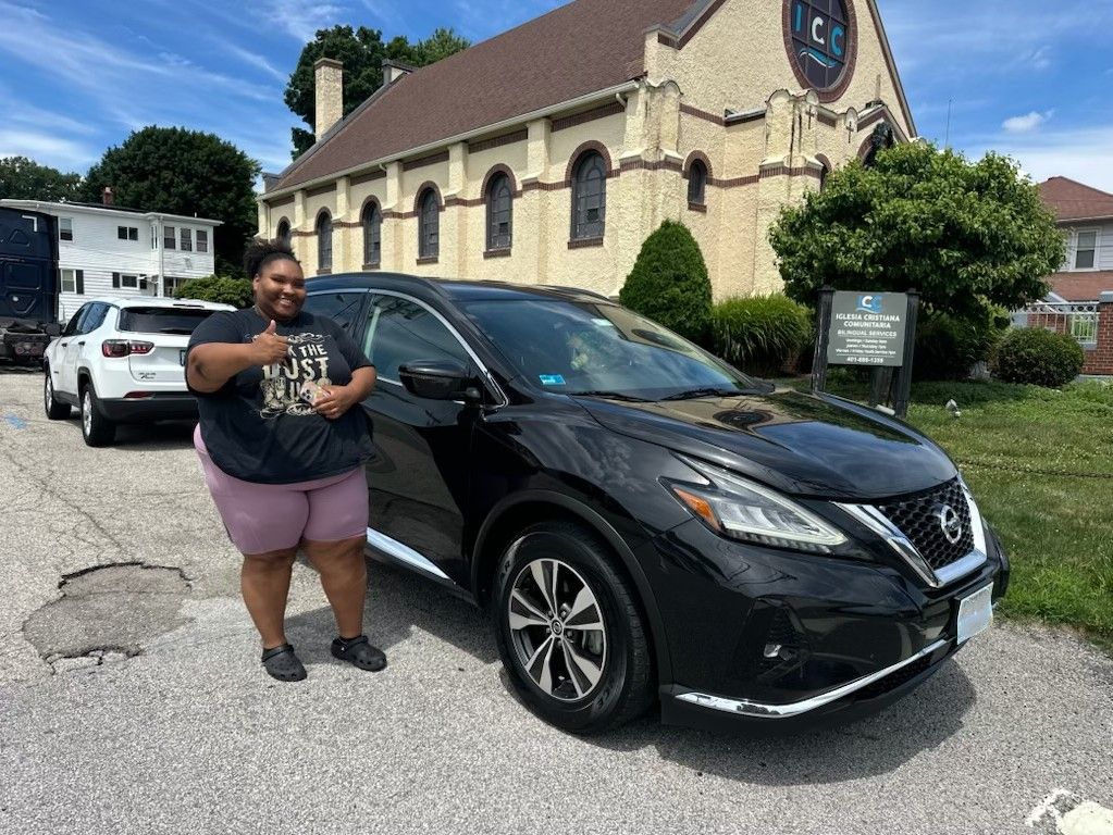 Person standing beside a black SUV in a parking lot near a stone building.