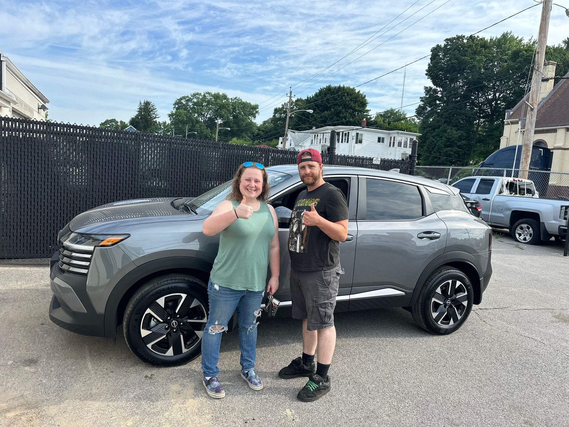 Two people standing and smiling beside a gray SUV in a parking lot, with houses and trees behind them