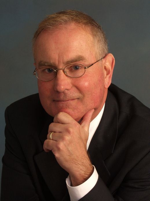 Man in suit, glasses, and ring, hand on chin, looking towards viewer.