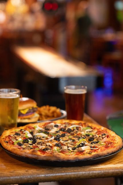 Pizza, burger, and drinks on a wooden table in a warmly lit restaurant or bar.