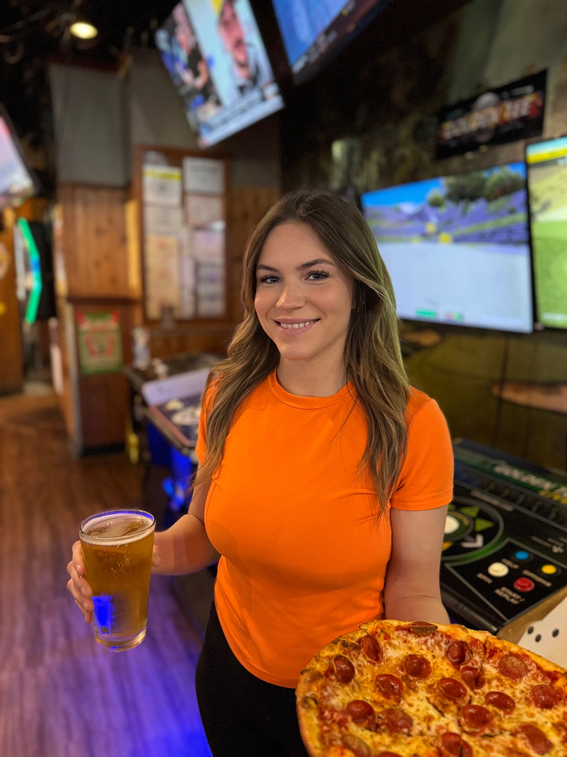 A Rooster's Bar & Grill Team member holding a drink and a pizza slice in a sports bar with TVs in the background