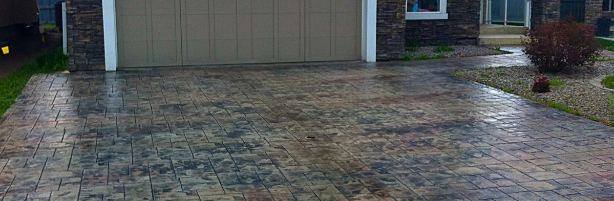 A wet brick driveway leading to a garage door. A small bush sits to the right, and grass borders.