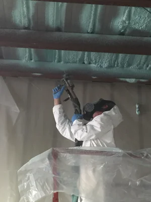 Person in protective gear spraying insulation on a ceiling, indoors.