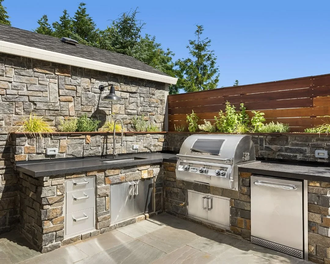 Outdoor kitchen with stone facade, stainless steel grill, and cabinets. Sunny day.