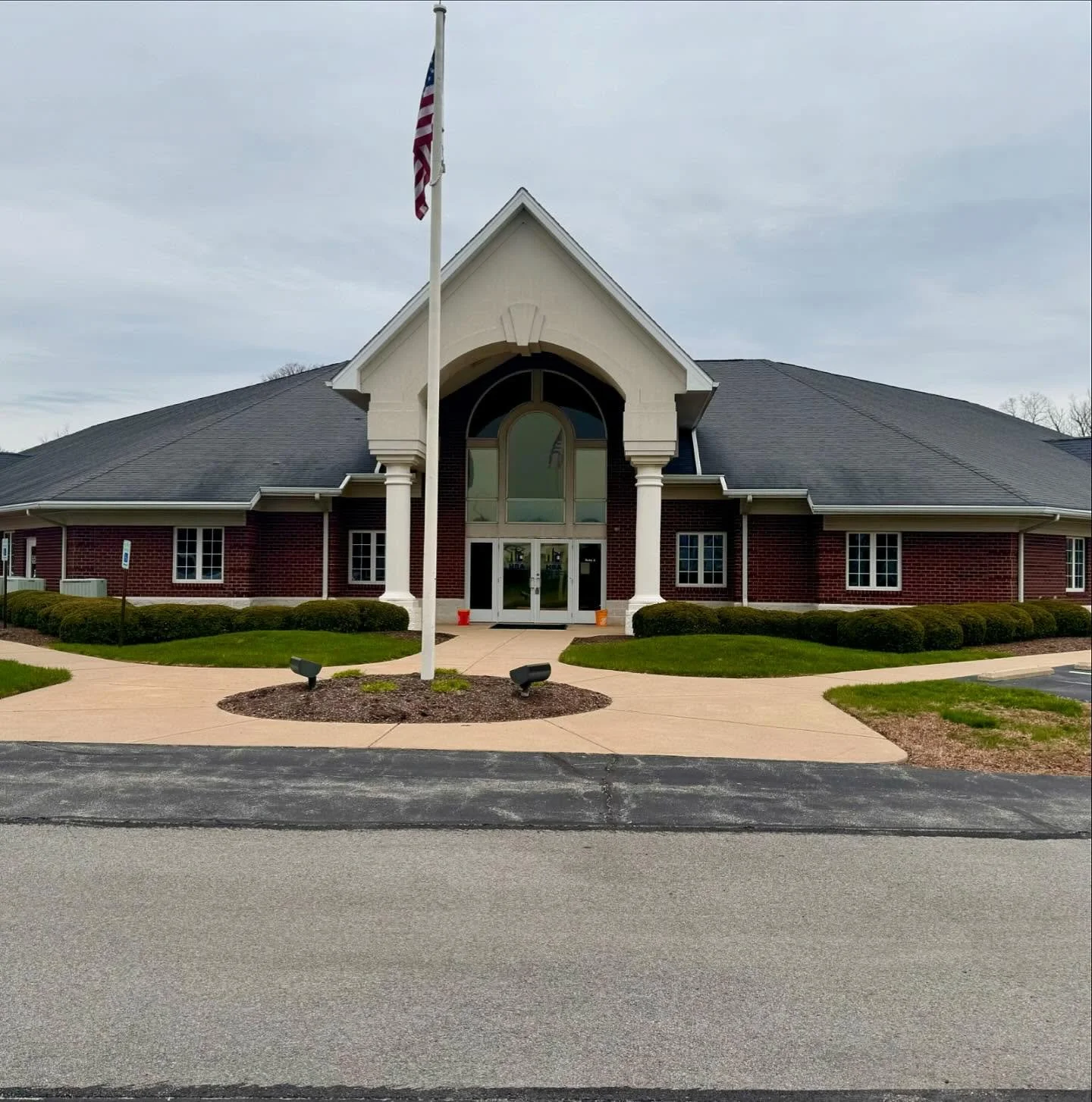 Brick building with white pillars, arched entryway, flag pole, and landscaped lawn on a cloudy day.