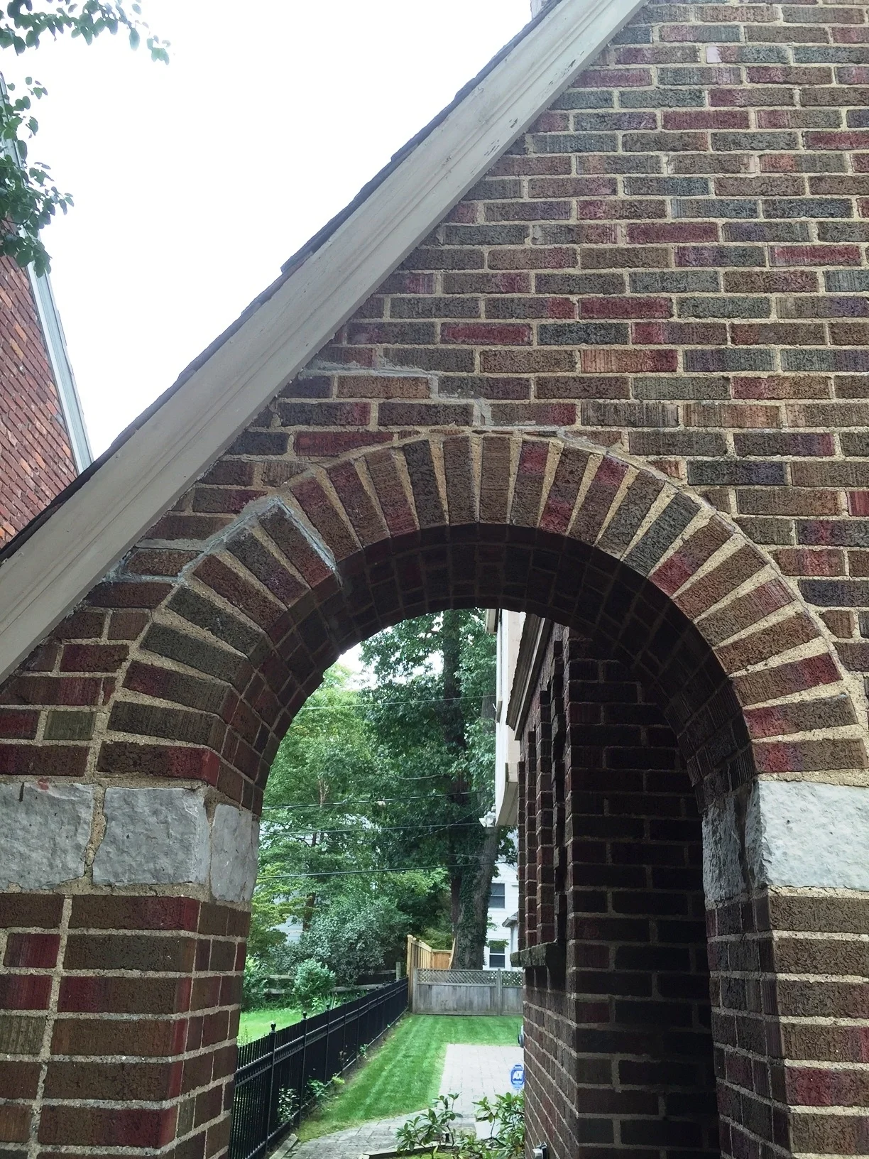 Brick archway in a brick wall, with a glimpse of a green yard and trees.