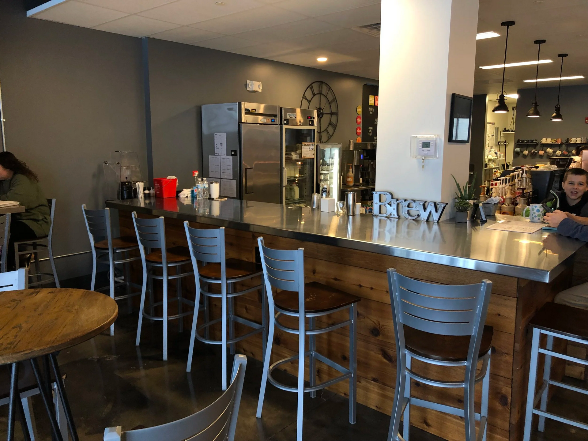 Coffee shop interior with a bar and seating; gray walls, stainless steel counter, wooden accents.