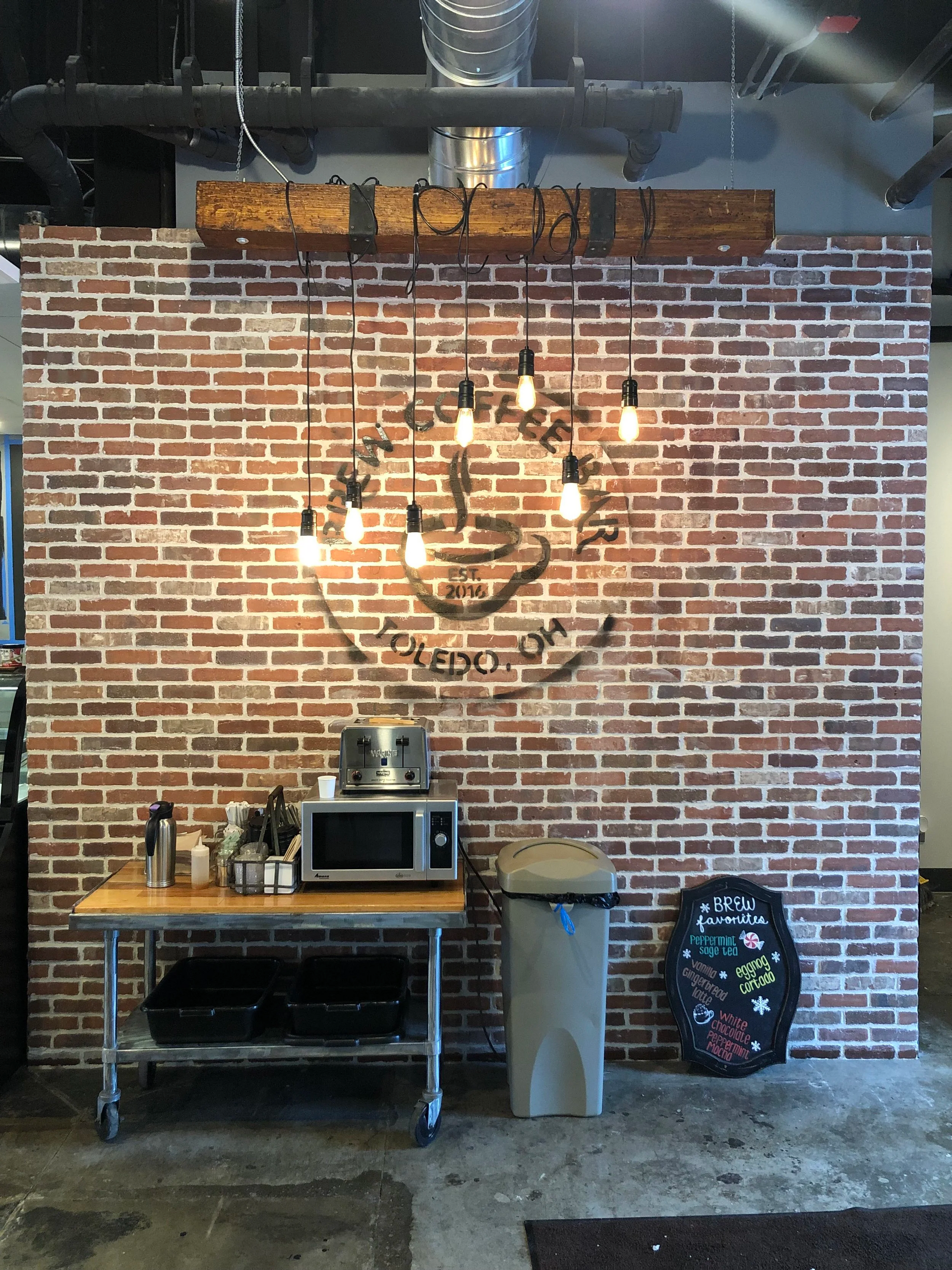 Brick wall with a metal-framed table holding a microwave and supplies. Overhead, a chandelier is mounted on a wooden beam.
