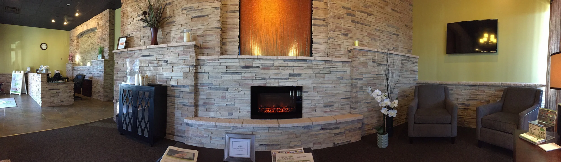 Interior waiting room with stone fireplace, armchairs, and TV on a light green wall.