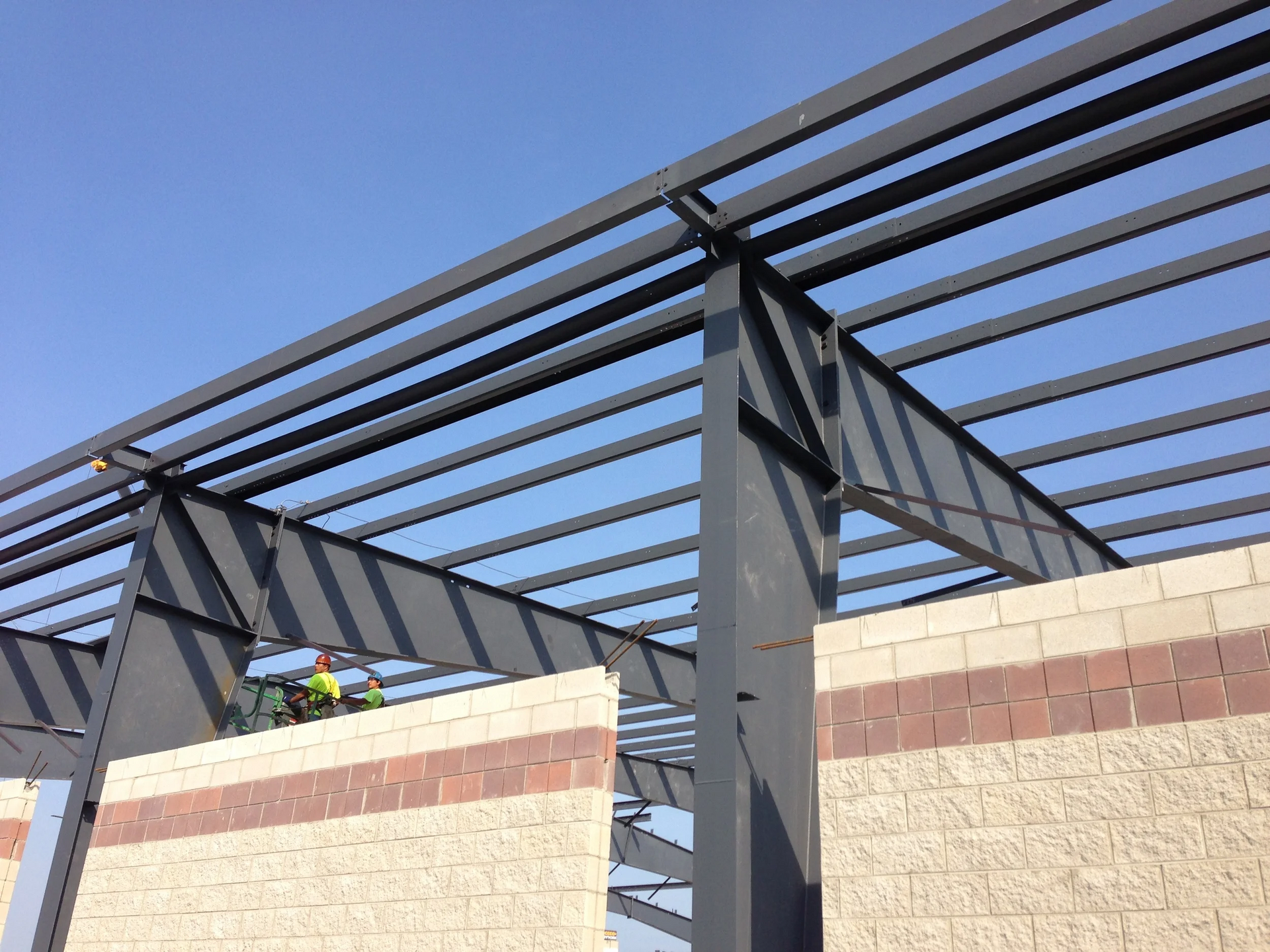 Steel beams of a building under construction, atop tan and red brick walls, under a clear blue sky.